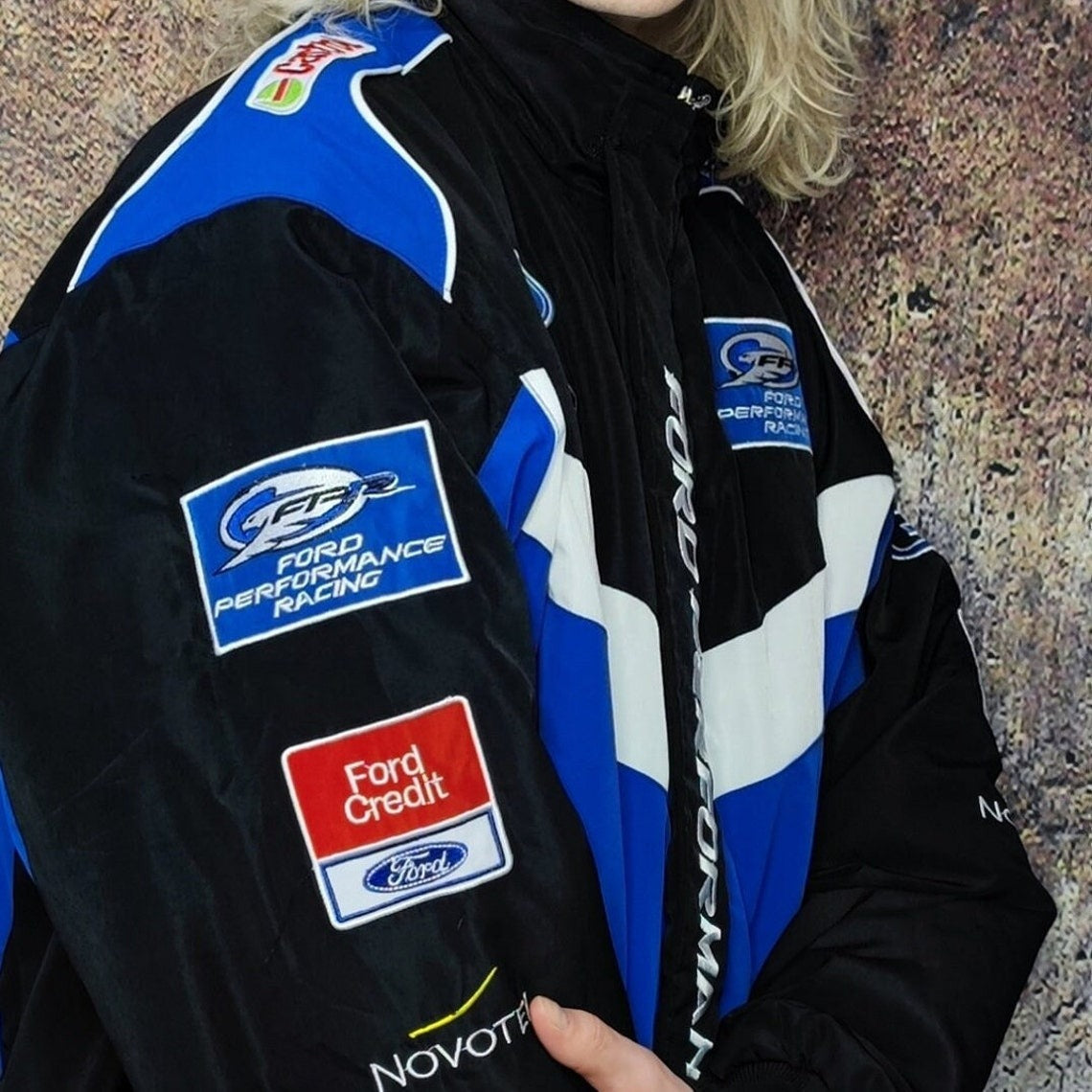 A person wears the Ford Vintage Jacket - Performance Edition in black, blue, and white, featuring patches that read “Ford Credit,” “Novotel,” and “Ford Performance Racing.” Only part of their face and blonde hair is visible.