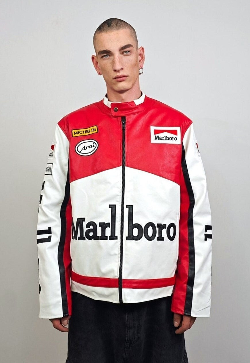 A young person with a buzz cut and hoop earring wears the Marlboro Racing Leather Jacket, a retro red, white, and black piece with bold Marlboro branding and sponsor logos, standing against a plain light background.