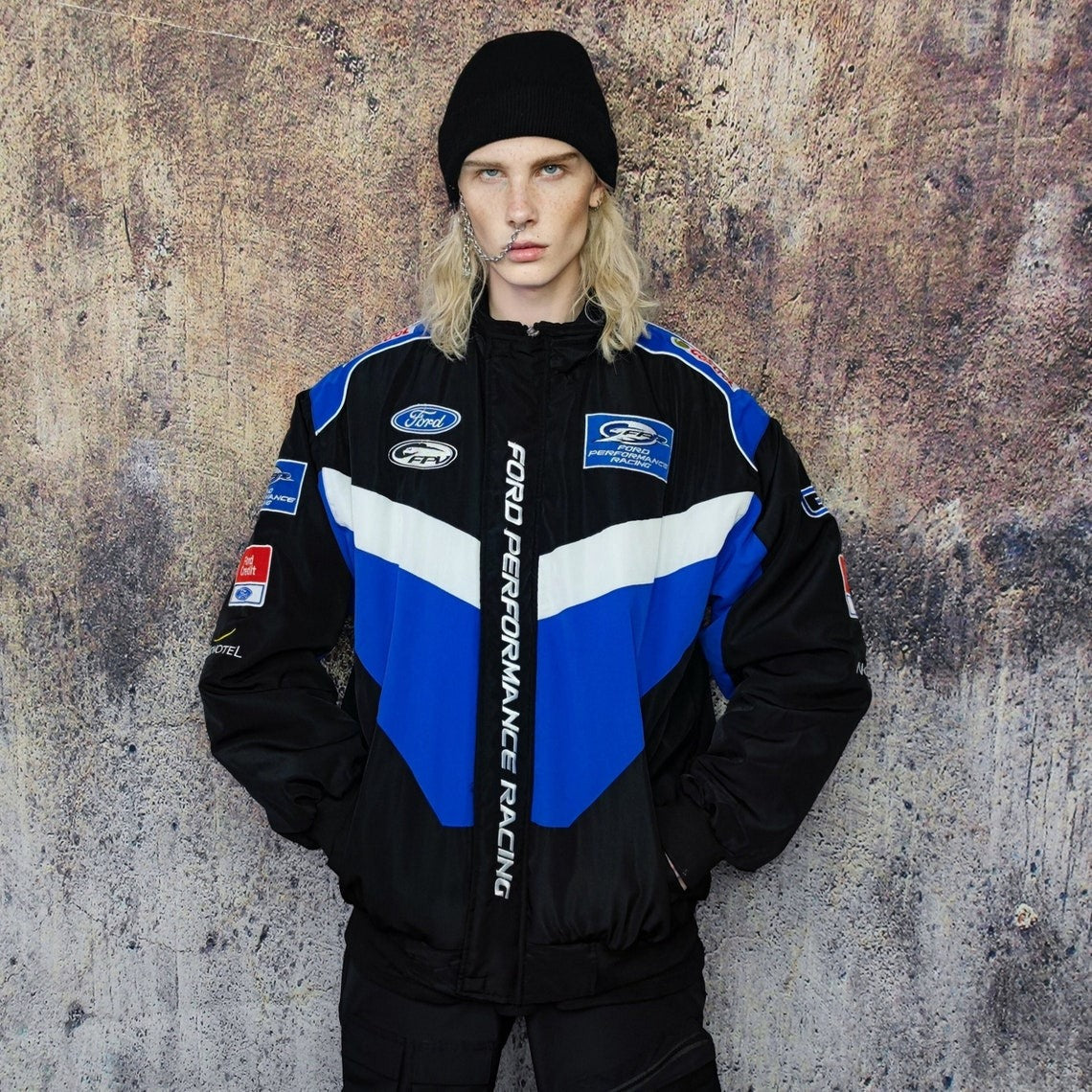 A person with long blonde hair, wearing a black beanie and the Ford Vintage Jacket - Performance Edition in blue, black, and white, stands against a textured brown wall.