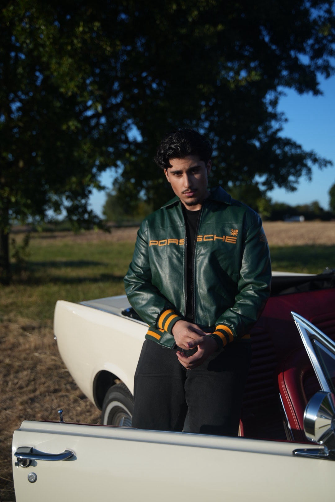 A man wearing the Porsche Vintage Leather Bomber Jacket - Green Edition stands next to an open car door, with a classic white convertible behind him and trees in the background on a sunny day, embodying true motorsport heritage.