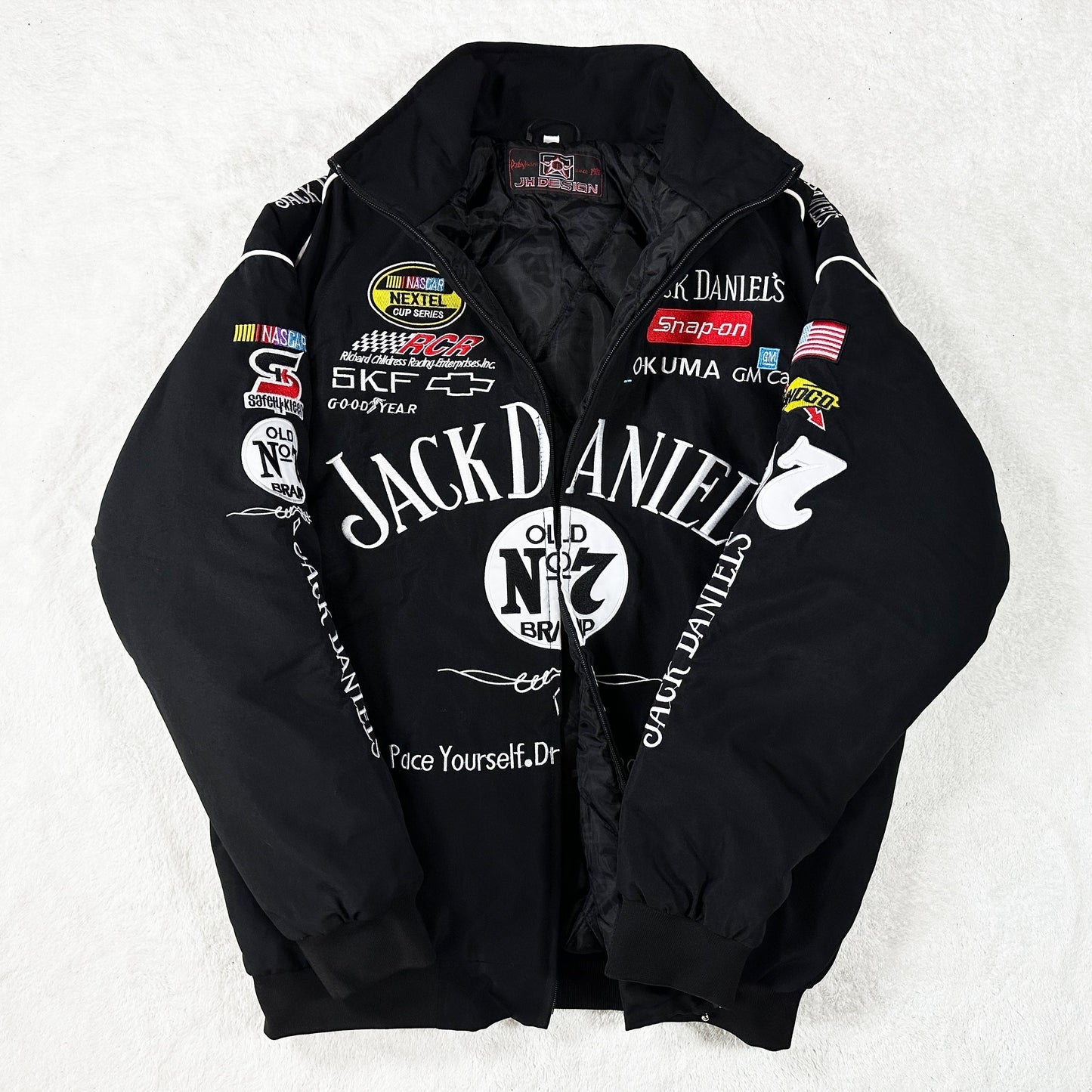 The Jack Daniel’s Racing Vintage Jacket, inspired by classic motorsport style, showcases sponsor patches and white embroidered logos on the front, sleeves, and collar—all displayed flat on a white surface.