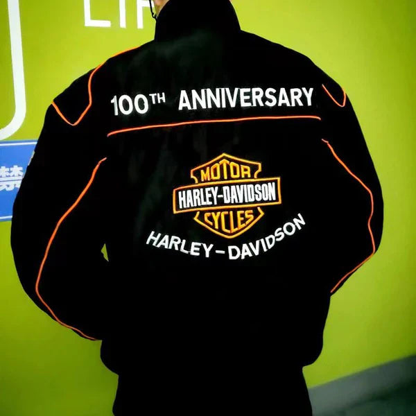 A person wears the Vintage Harley Davidson Jacket with orange trim and 100th Anniversary branding, standing against a green background and capturing timeless retro motorcycle style.