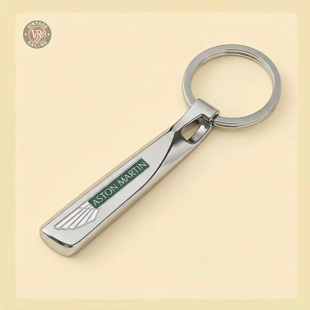 The Aston Martin Keychain is a silver metal accessory with a green and white logo, featuring a rounded rectangular design and a keyring at the top, shown resting on a light beige surface.