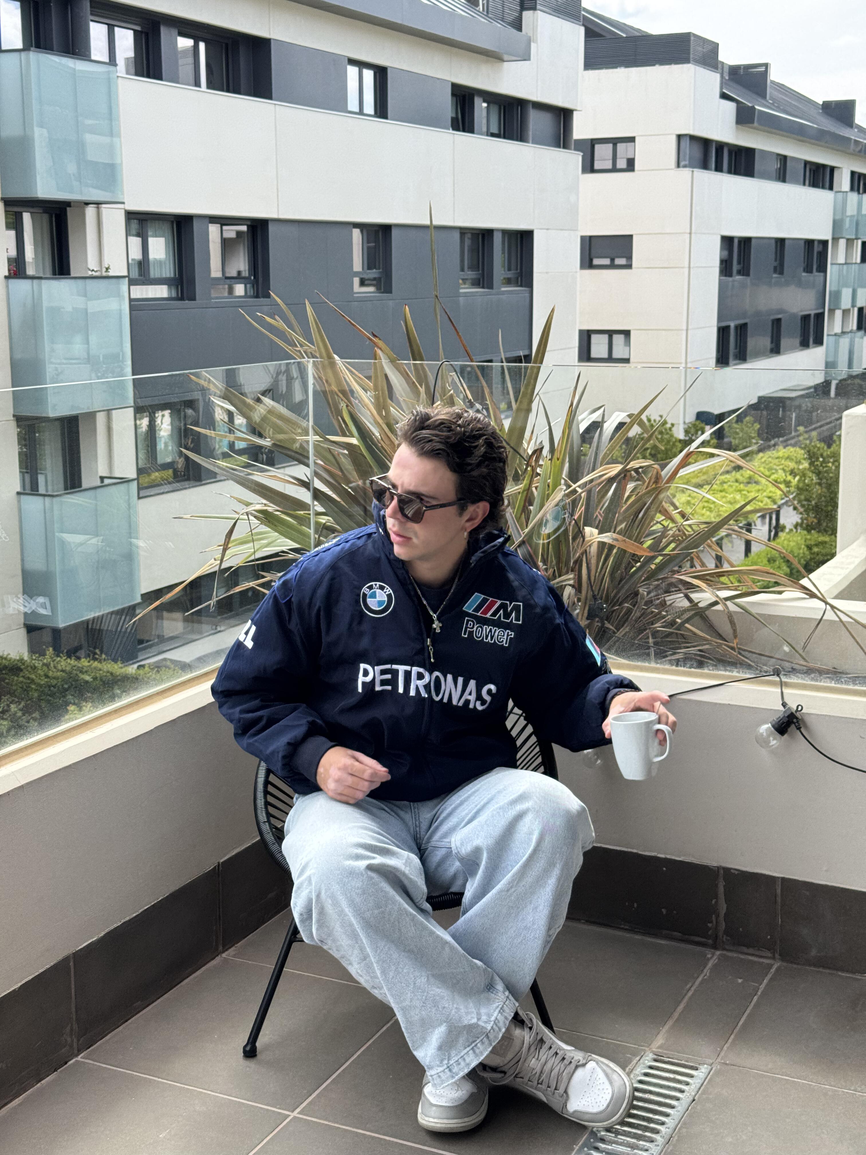 Wearing a navy BMW F1 Vintage Racing Jacket - Classic Edition and light jeans, a person with sunglasses sits on a balcony chair, holding a white mug and glancing sideways, with modern buildings and greenery behind them.