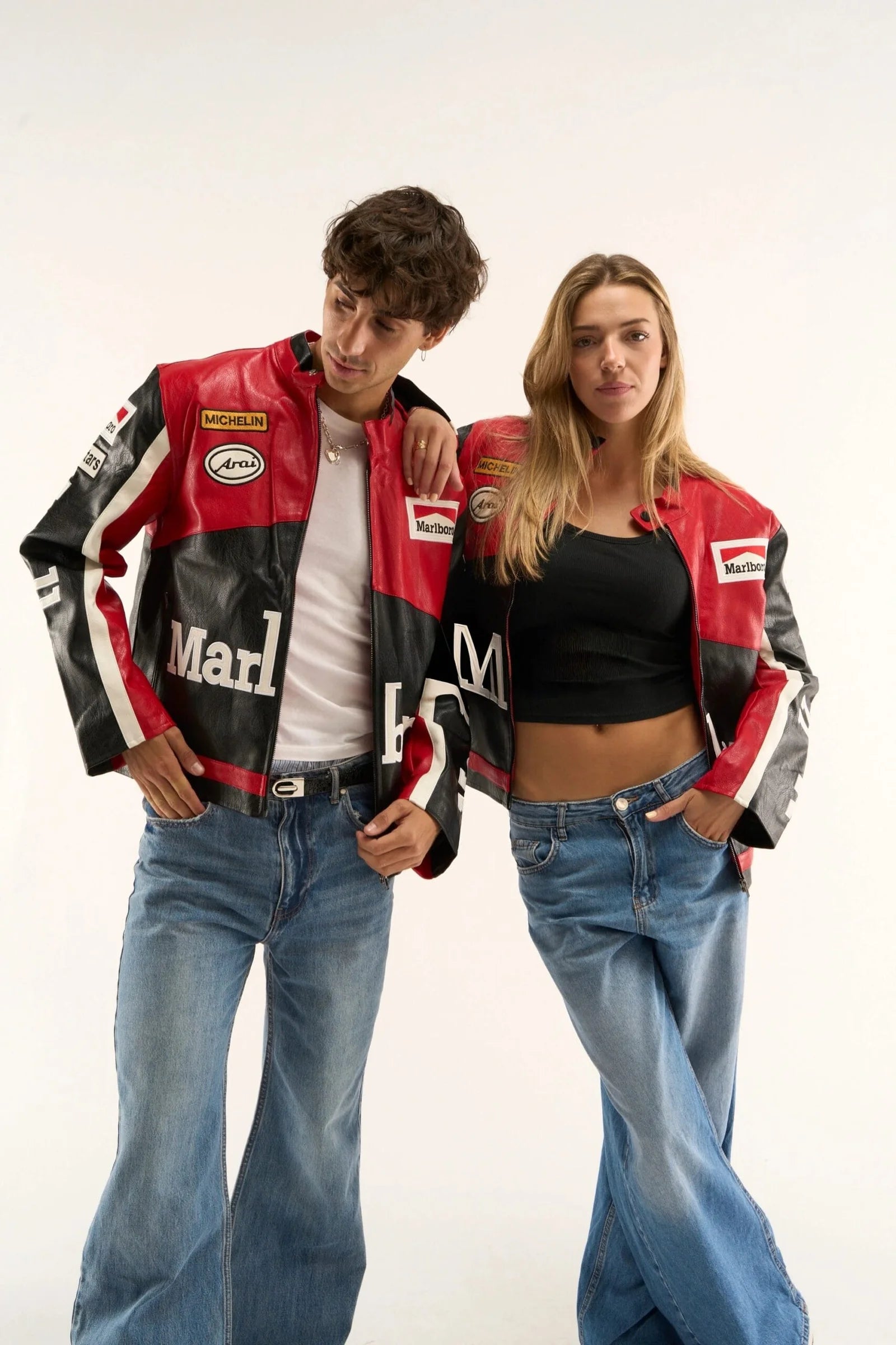 Two people stand together wearing matching Marlboro Racing Leather Jacket - Black Edition pieces, paired with white and black tops and blue jeans. They pose confidently against a plain white background.