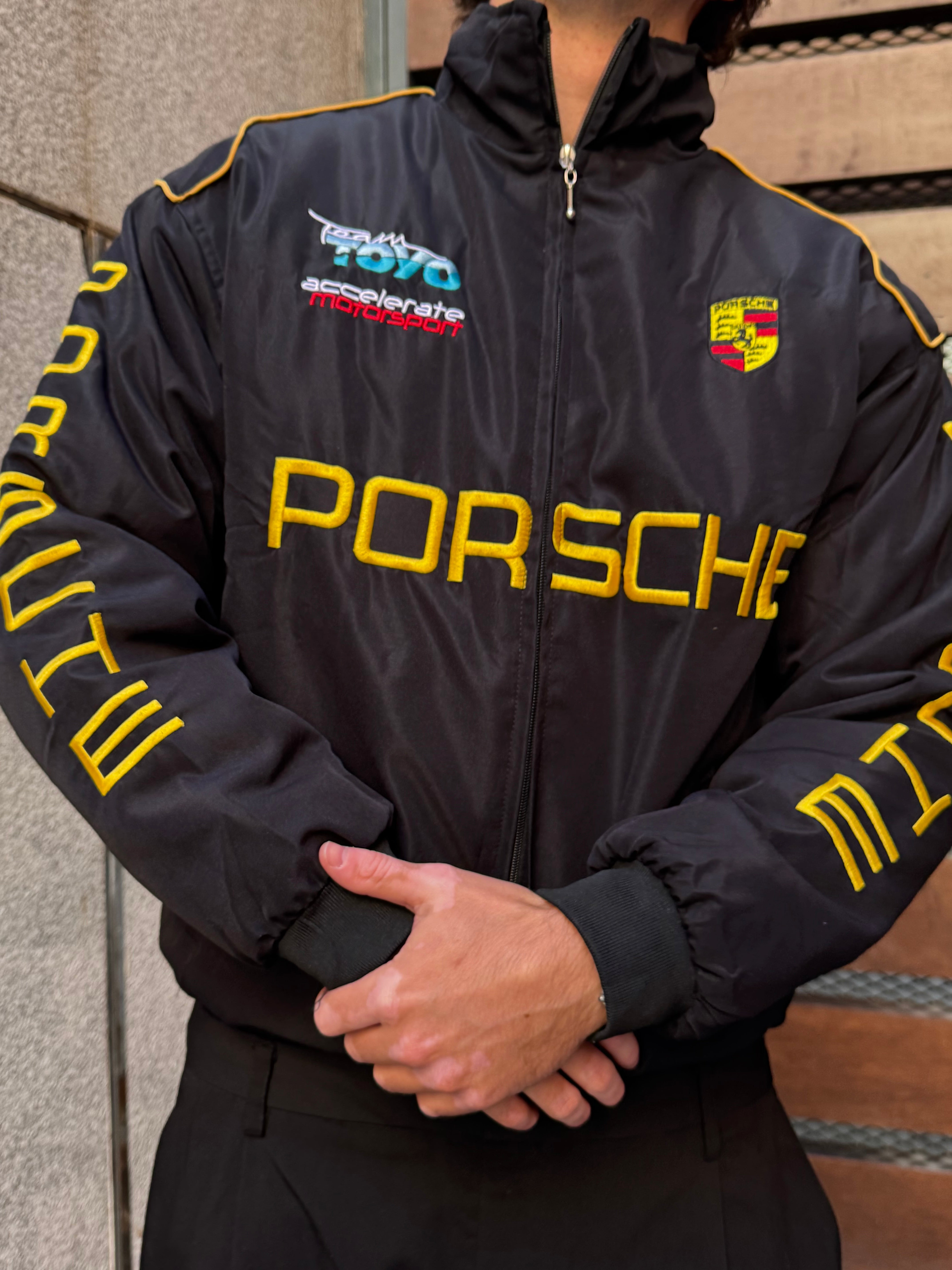 Wearing a Porsche Vintage Racing Jacket with yellow lettering and embroidered racing logos, a person stands facing the camera with hands clasped in front. The background features a beige wall and a wooden door.