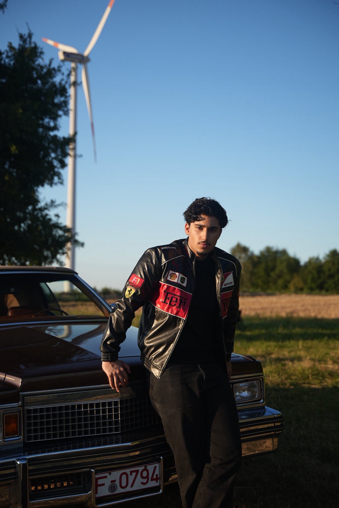 A young man wearing the Ferrari F1 Vintage Racing Leather Jacket – Red Edition leans against a vintage car in a field, with a wind turbine and trees in the background beneath a clear blue sky.