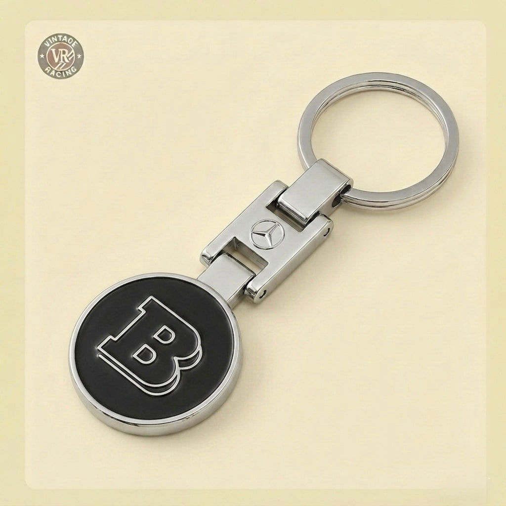 The Brabus Keychain features a silver design with a round black pendant, showcasing a raised "B" at its center on a metallic key ring. Ideal for fans of the celebrated Mercedes-Benz tuner. Displayed against a light beige background.