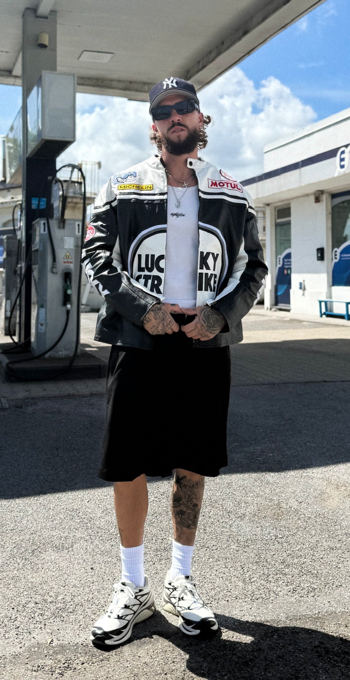A man in sunglasses, a black cap, and the Lucky Strike Vintage Racing Leather Jacket - White & Black Edition stands at a gas station. He wears a white shirt, black shorts, white socks, and sneakers, with visible tattoos on his arms and legs.