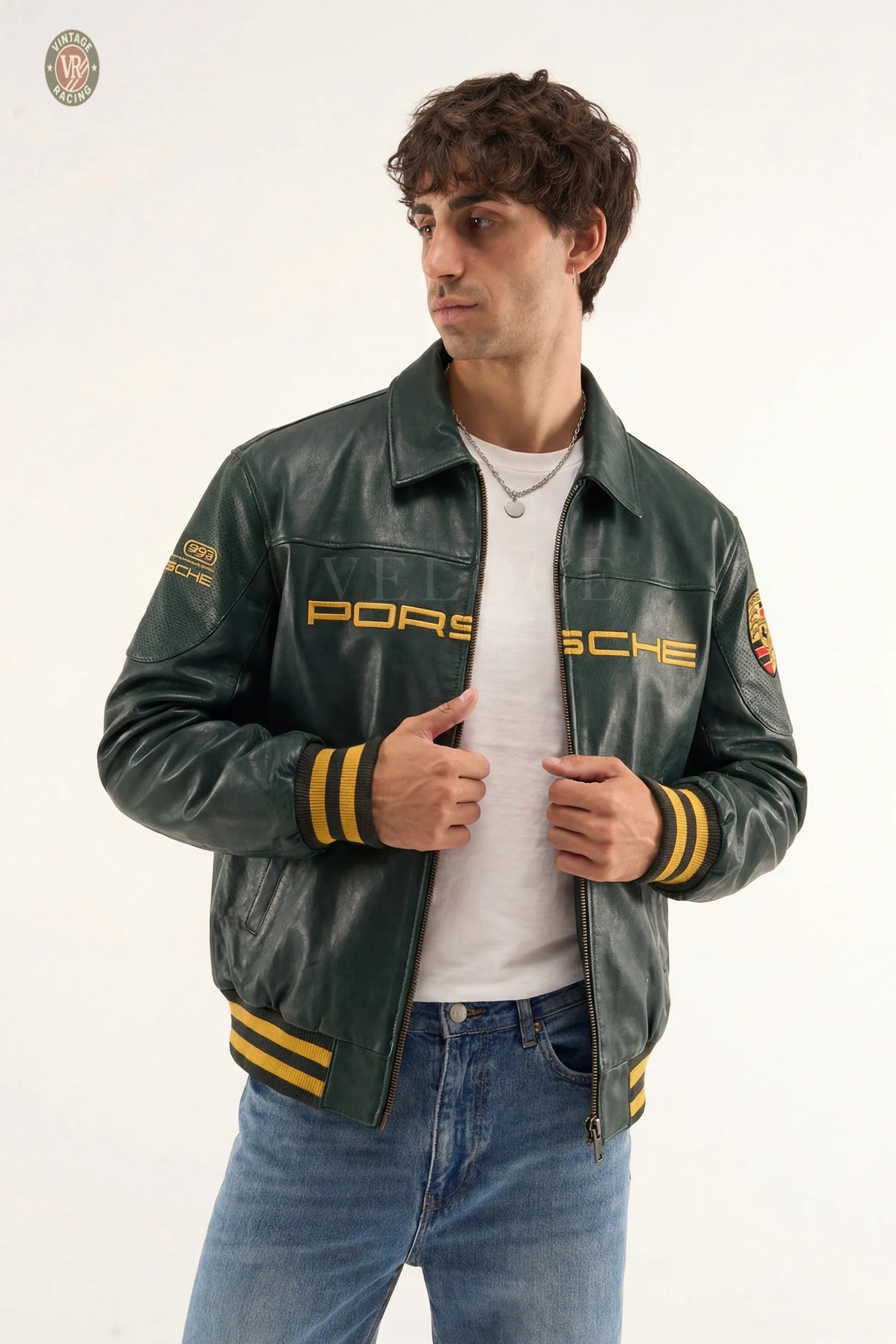 A man in a Porsche Vintage Leather Bomber Jacket - Green Edition with yellow striped sleeves and waistband, over a white t-shirt and blue jeans, stands against a plain white background, evoking classic motorsport heritage.