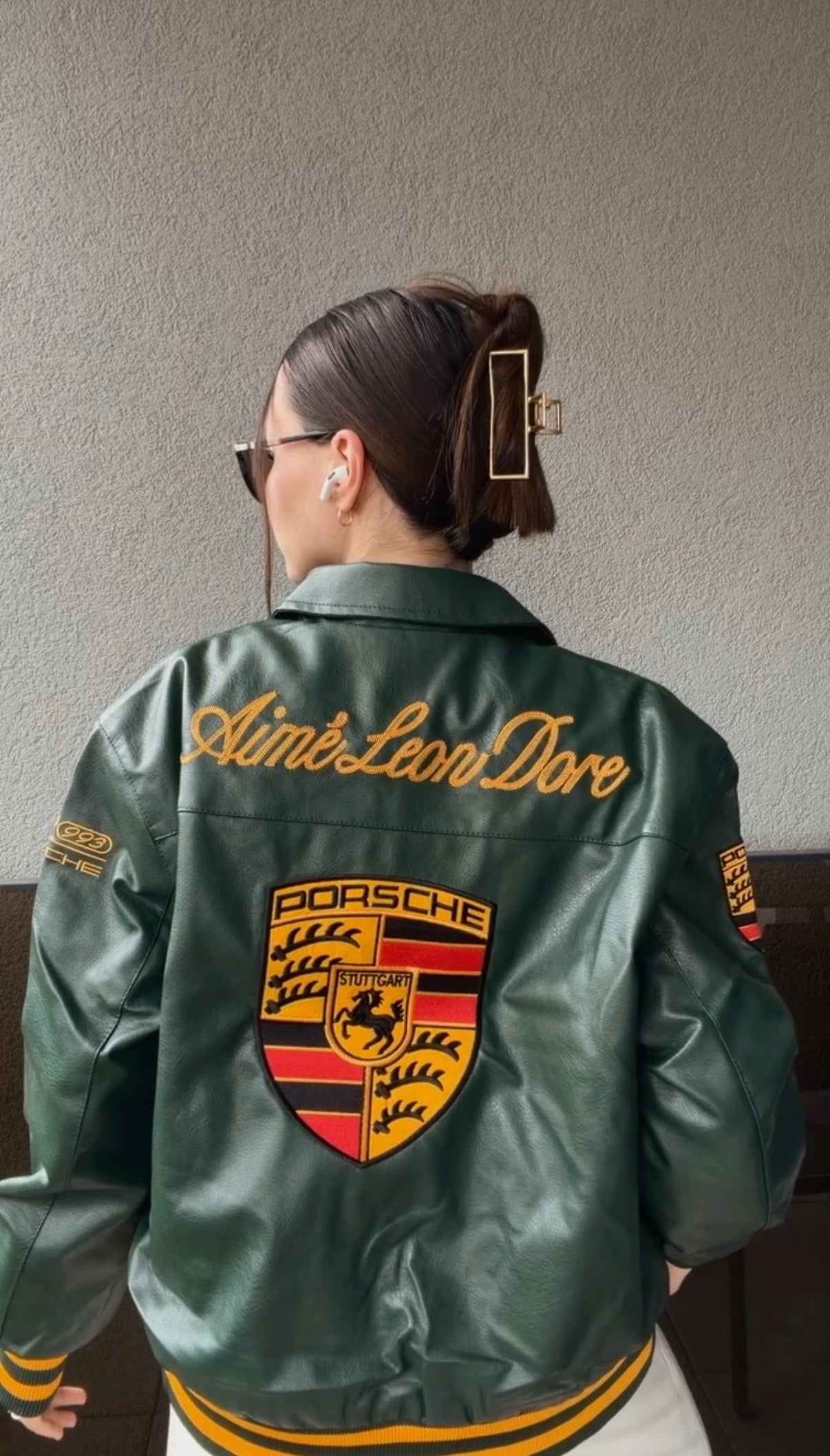 A woman with sunglasses and a large hair clip faces away, wearing the Porsche Vintage Leather Bomber Jacket - Green Edition with yellow Aimé Leon Dore text and a bold Porsche crest. She also has wireless earbuds in her ears.