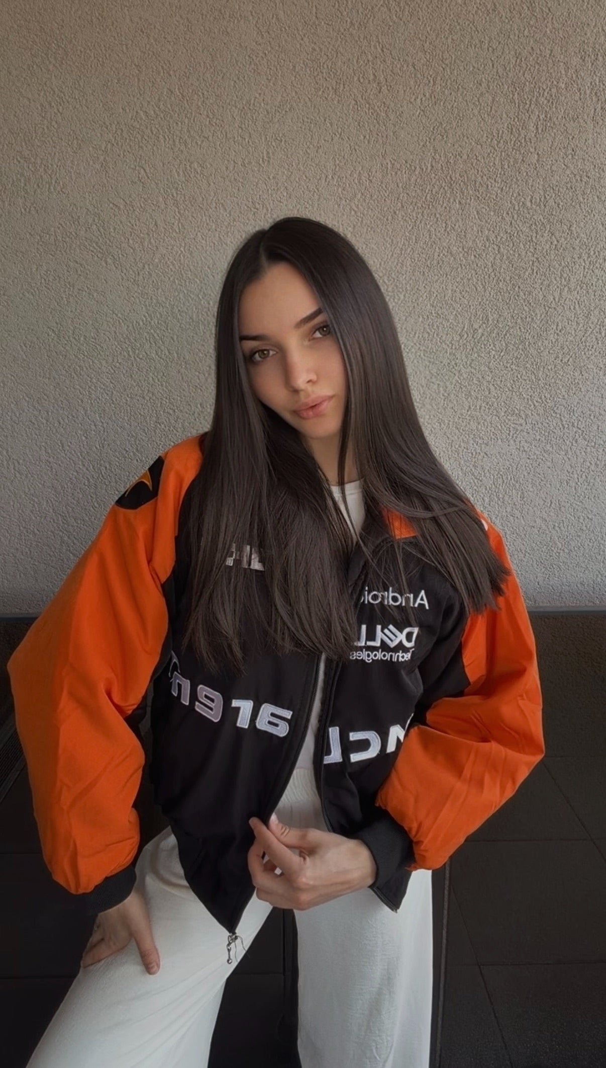 A woman with long brown hair stands indoors, wearing a McLaren F1 Vintage Racing Jacket in striking orange and black over a white outfit, posing with one hand near her waist against a light textured wall.