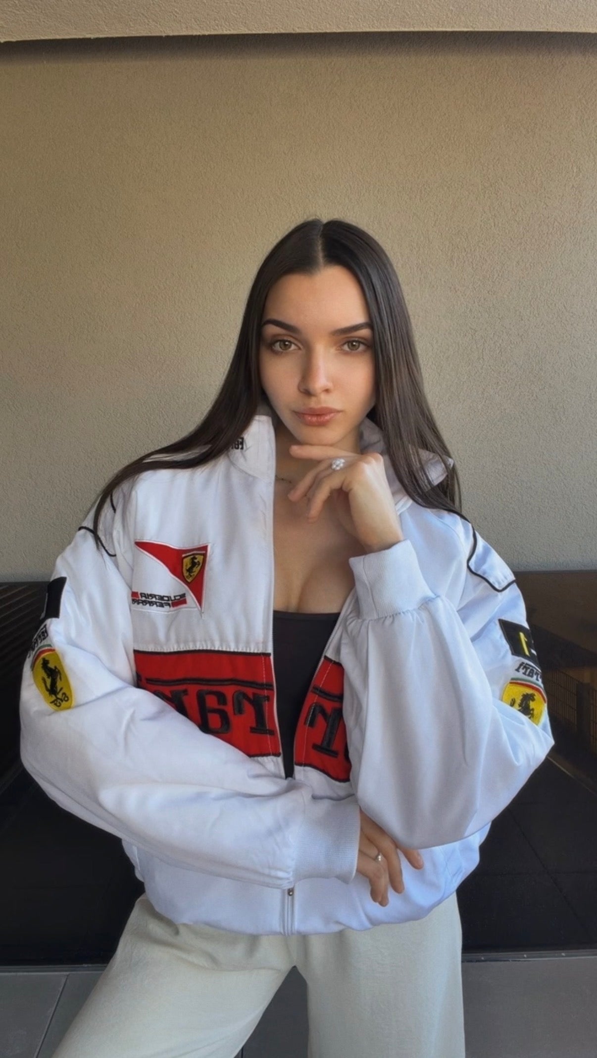 A woman with long brown hair is wearing the Ferrari F1 Vintage Racing Jacket - White Edition, posing with one hand near her face while standing indoors against a neutral wall.