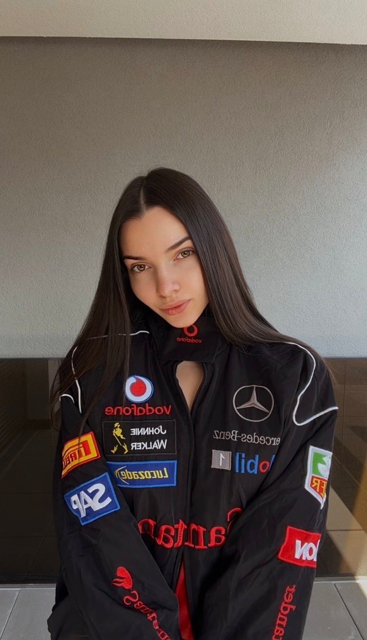 A woman with long brown hair wears the Mercedes-Benz Santander Vintage Jacket, featuring colorful sponsor logos, as she stands indoors against a plain gray wall and looks at the camera with a neutral expression that evokes motorsport heritage.