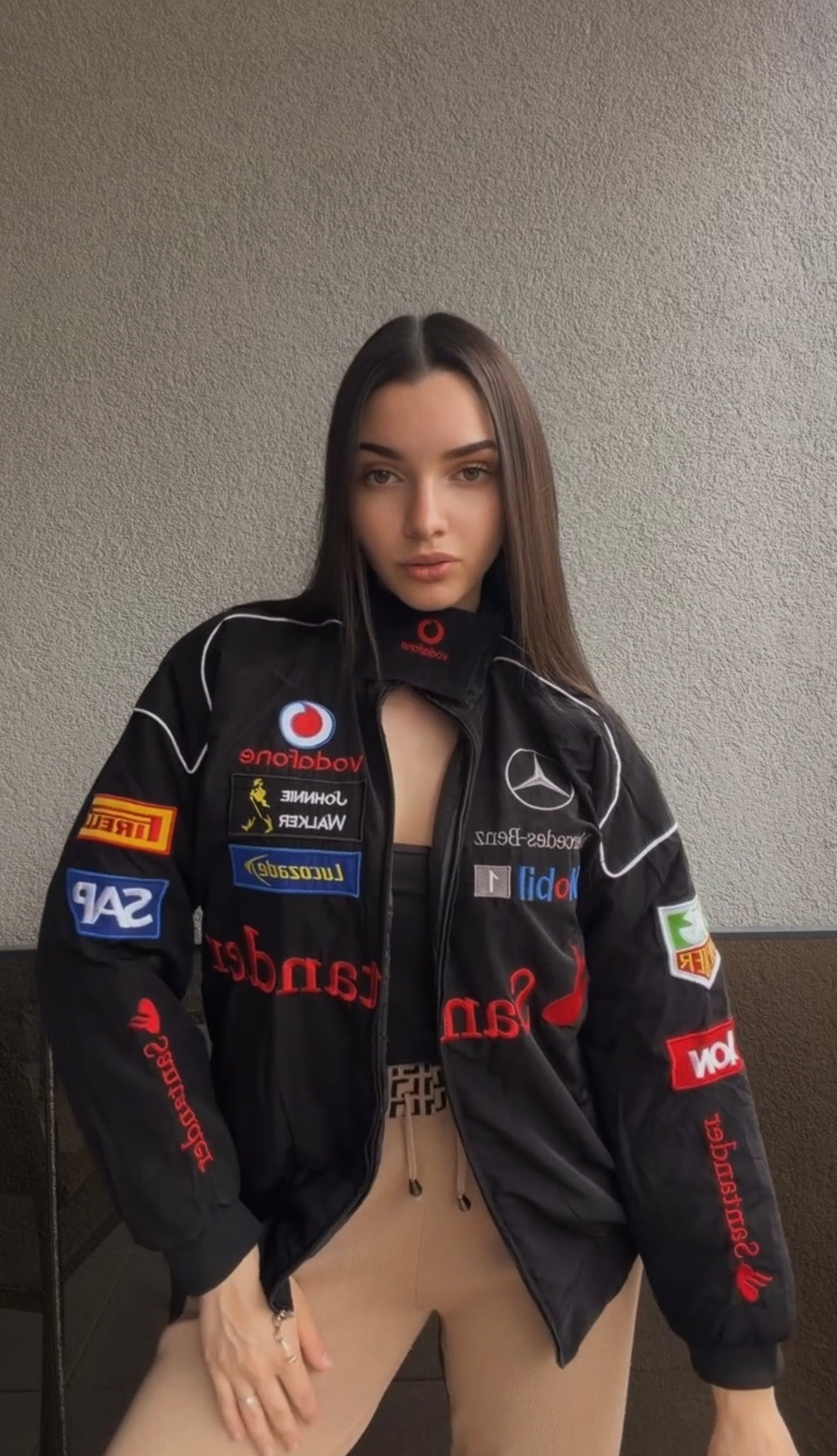 A woman with long brown hair wears the Mercedes-Benz Santander Vintage Jacket, which showcases a range of vibrant sponsor logos. She stands against a plain gray wall, capturing the essence of motorsport heritage and stealth racing style.