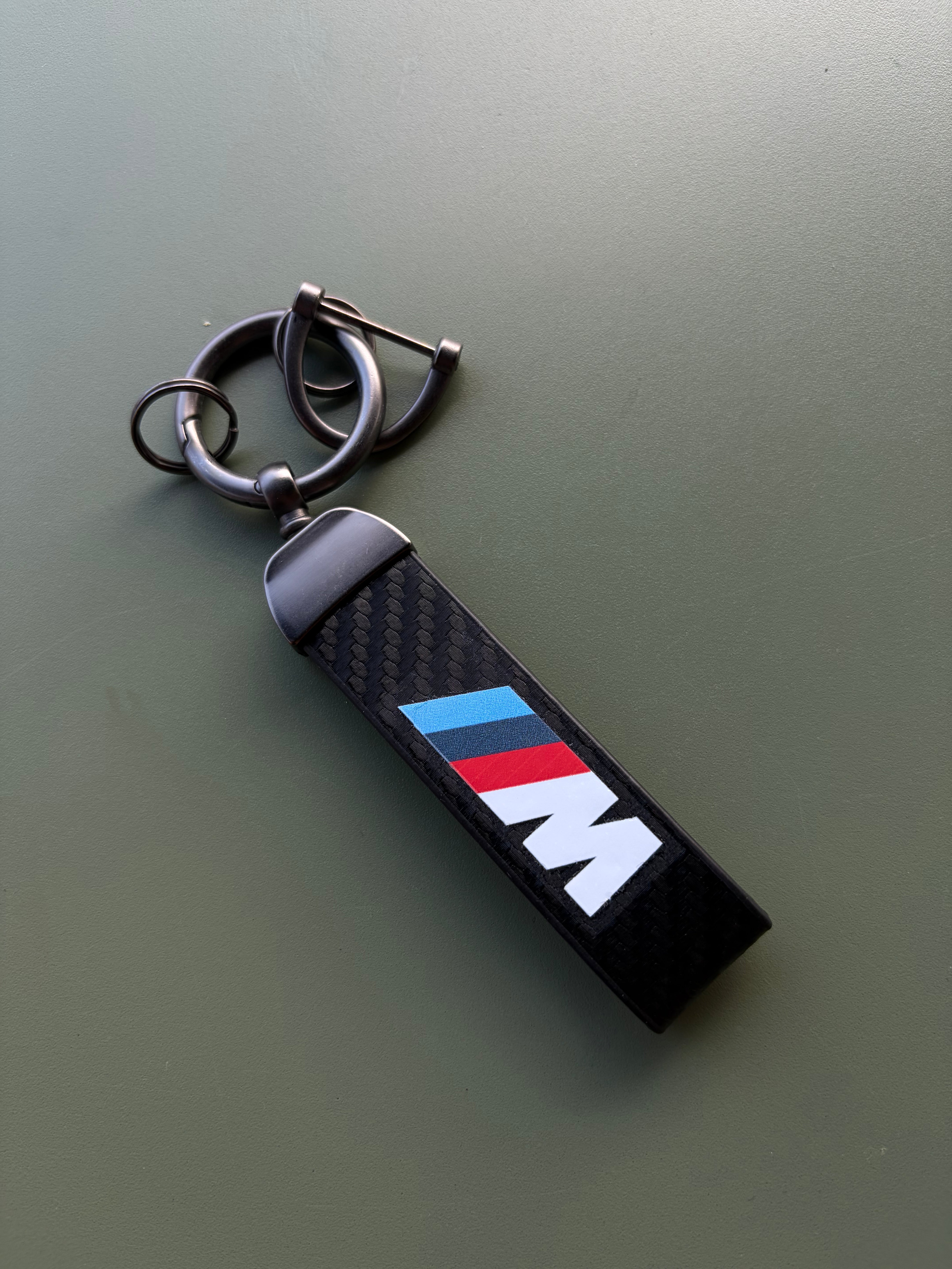 The BMW M Sport Keychain is a carbon fiber accessory with a metal ring, showcasing the iconic BMW M logo with blue, purple, and red stripes. Displayed on a green surface, it’s the ideal addition to your collection of M Sport accessories.