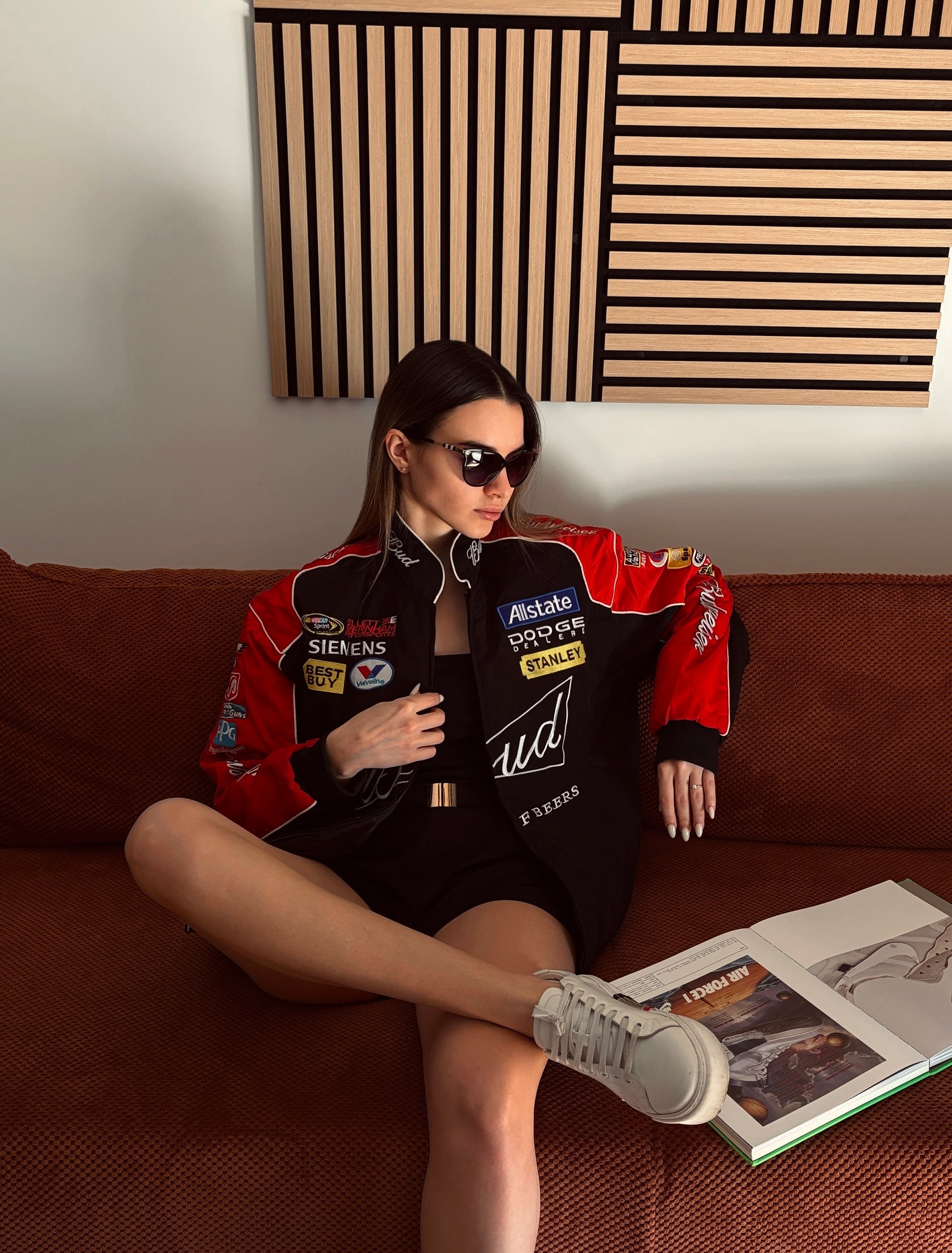 A woman in sunglasses and a Budweiser Vintage Racing Jacket - NASCAR Black Edition sits on a brown sofa with crossed legs, one arm resting on the backrest, and an open book on the cushion beside her. Abstract wood art hangs on the wall behind her.