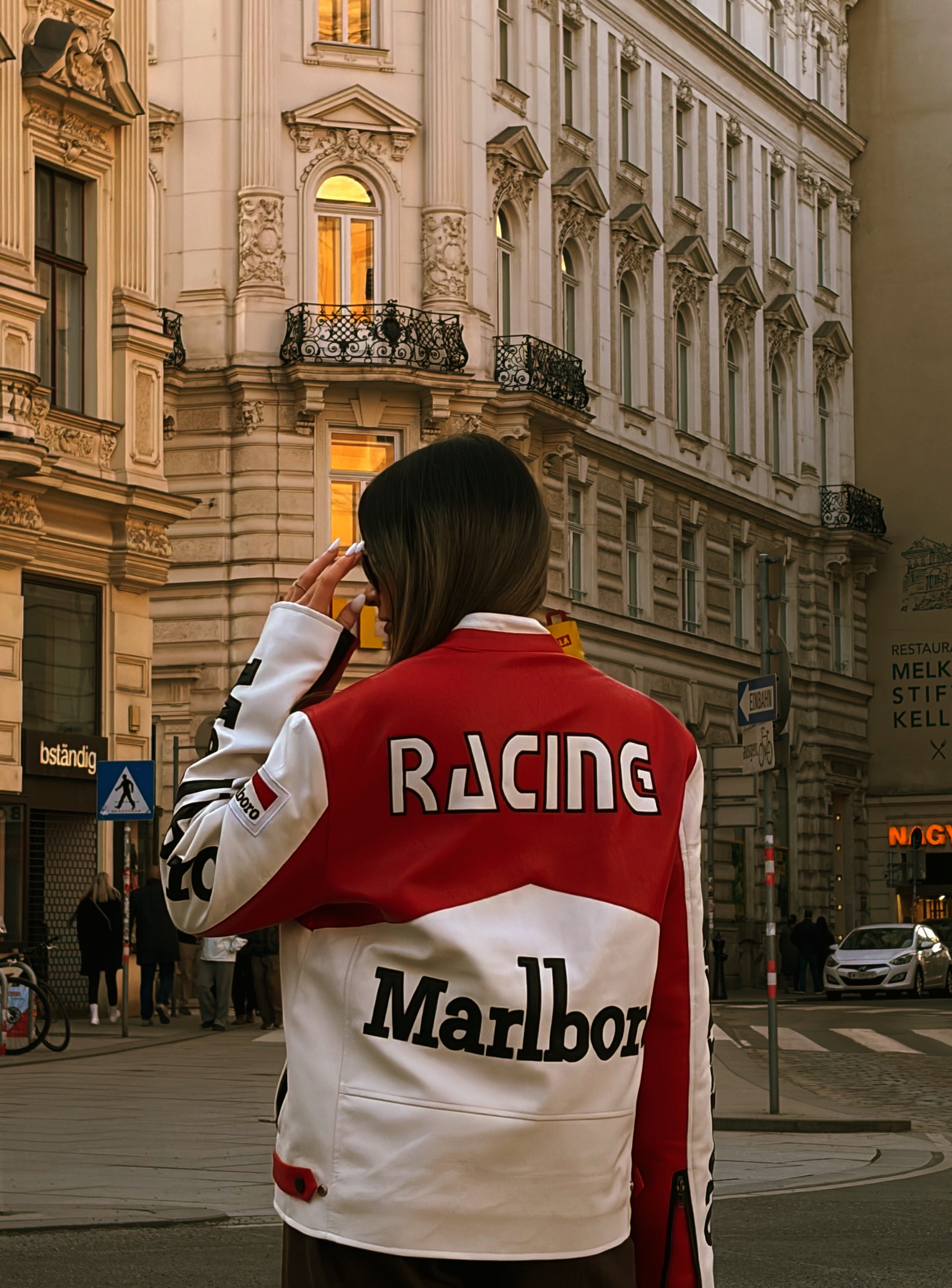 A person stands on a city street with their back to the camera, wearing the Marlboro Racing Leather Jacket. This jacket captures iconic retro motorsport style with bold “RACING” and “Marlboro” prints that evoke the adrenaline of 90s F1 racing, set against a backdrop of elegant historic buildings in the classic urban landscape.