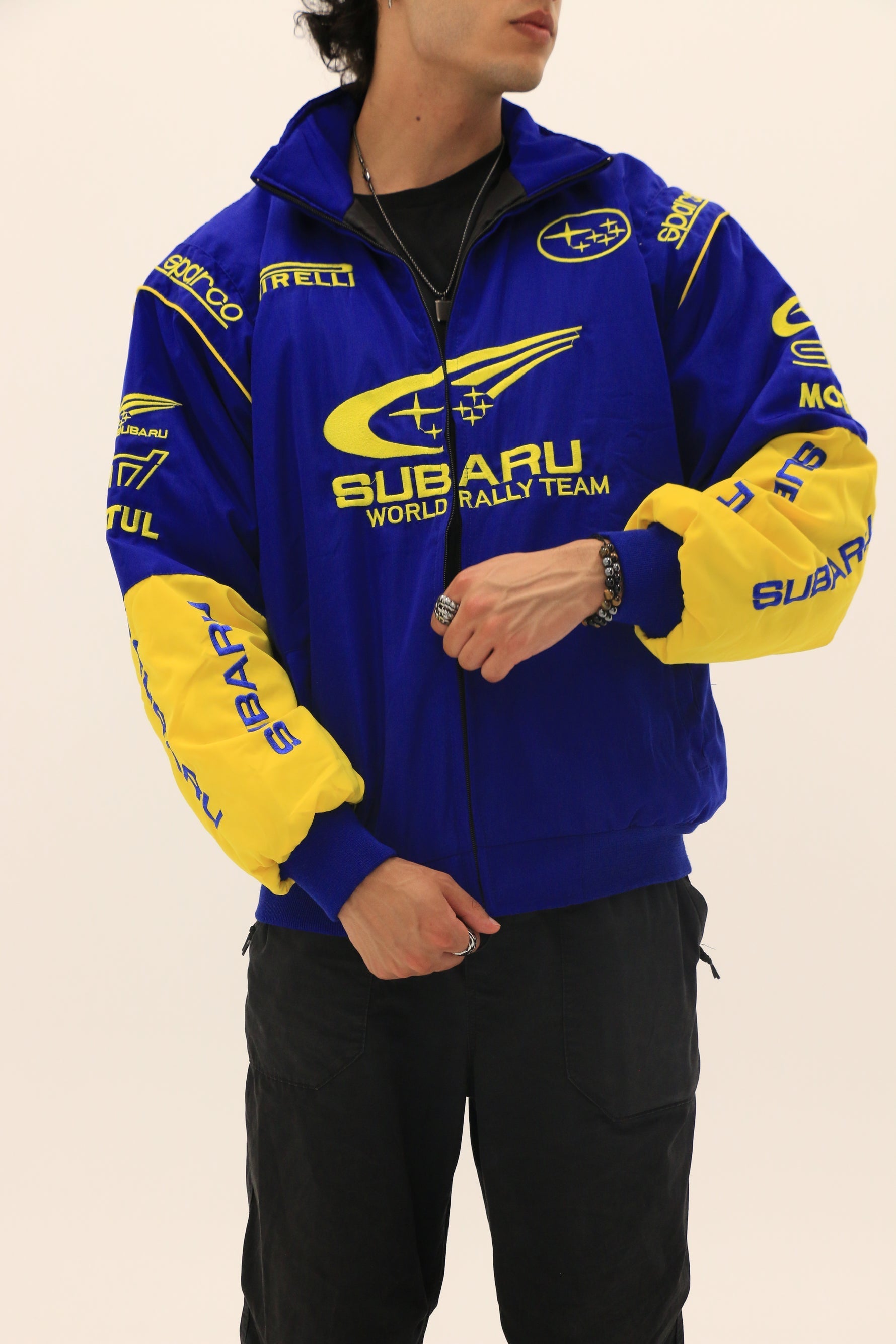 A person is wearing the Subaru World Rally Team Vintage Jacket with iconic blue and yellow 555 livery, paired with black pants, a necklace, and a watch, standing against a plain light background.