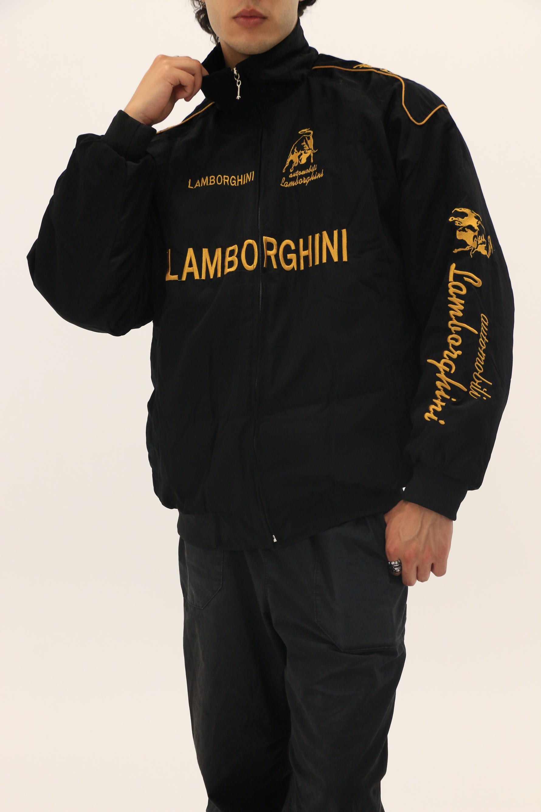 A person wears the Lamborghini Vintage Racing Jacket featuring yellow branding and logos on the chest, sleeves, and collar. Their face is partially obscured as they stand against a plain white background—ideal for motorsport fans.