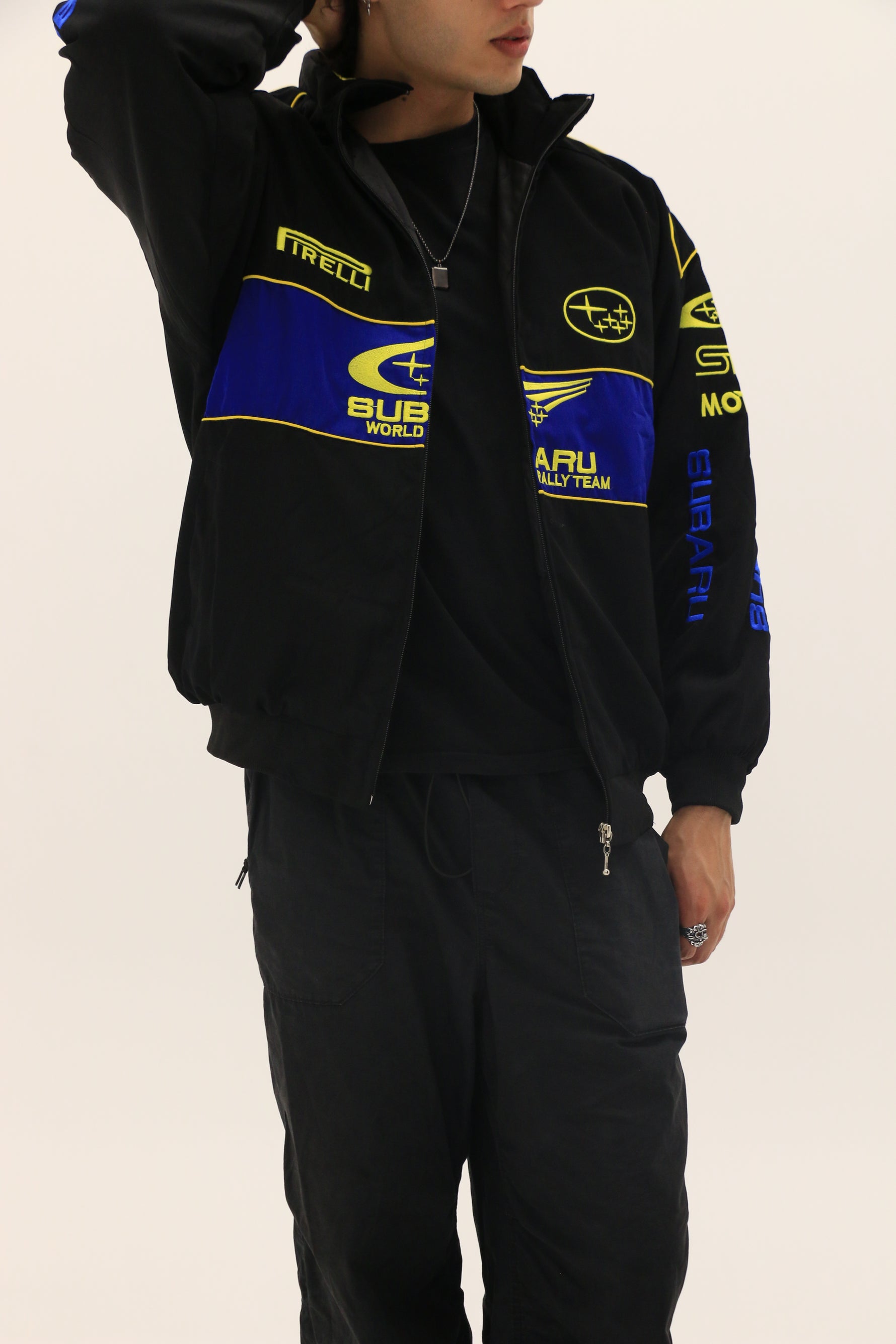 Wearing a Subaru Rally Vintage Racing Jacket in black and blue with yellow logos, paired with black pants and a silver ring, a person stands against a plain light background. Their face is partly out of frame, echoing rally heritage style.