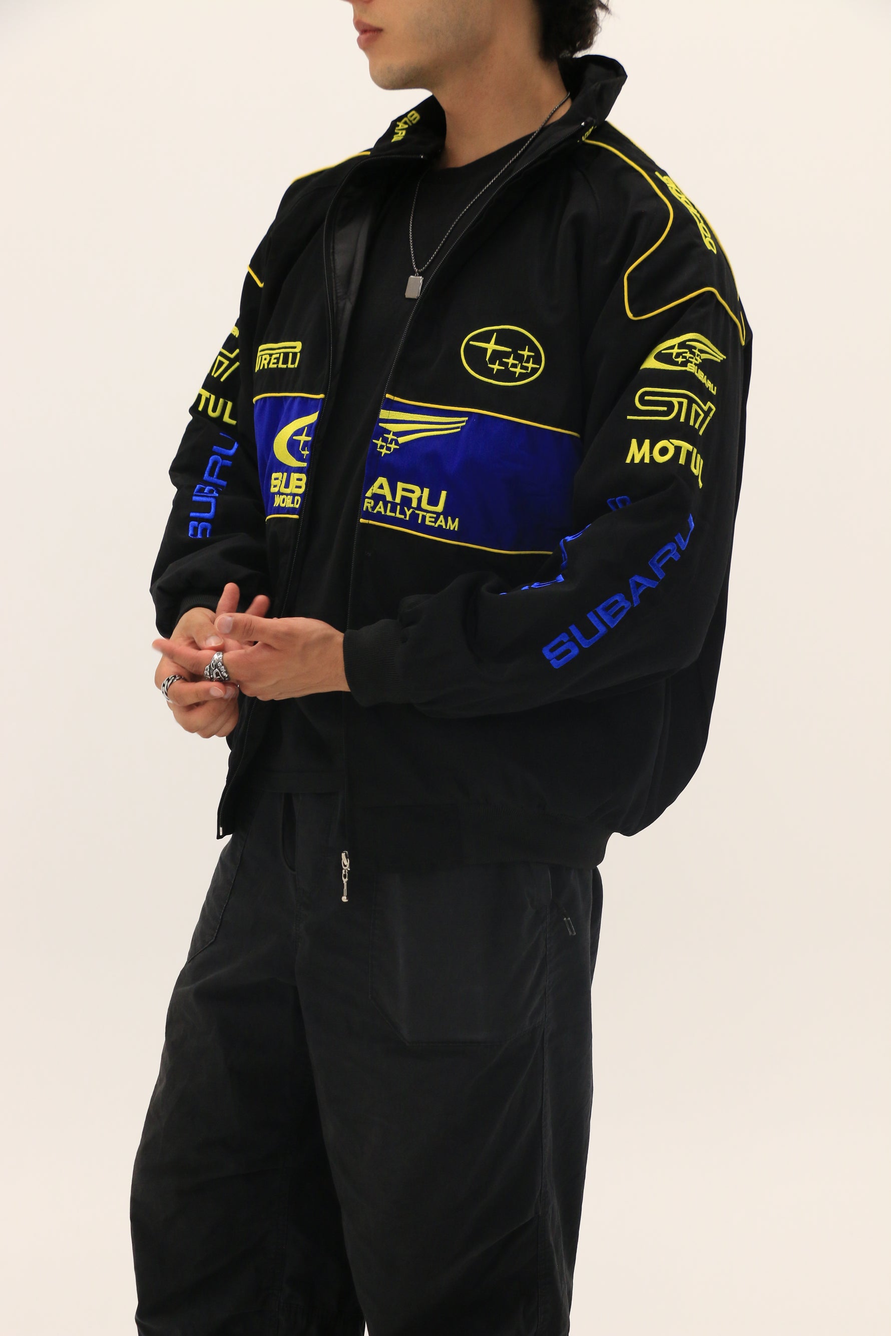 A person wears a Subaru Rally Vintage Racing Jacket—black with blue and yellow accents and sponsor logos—showcasing rally heritage, standing against a plain white background with hands clasped.