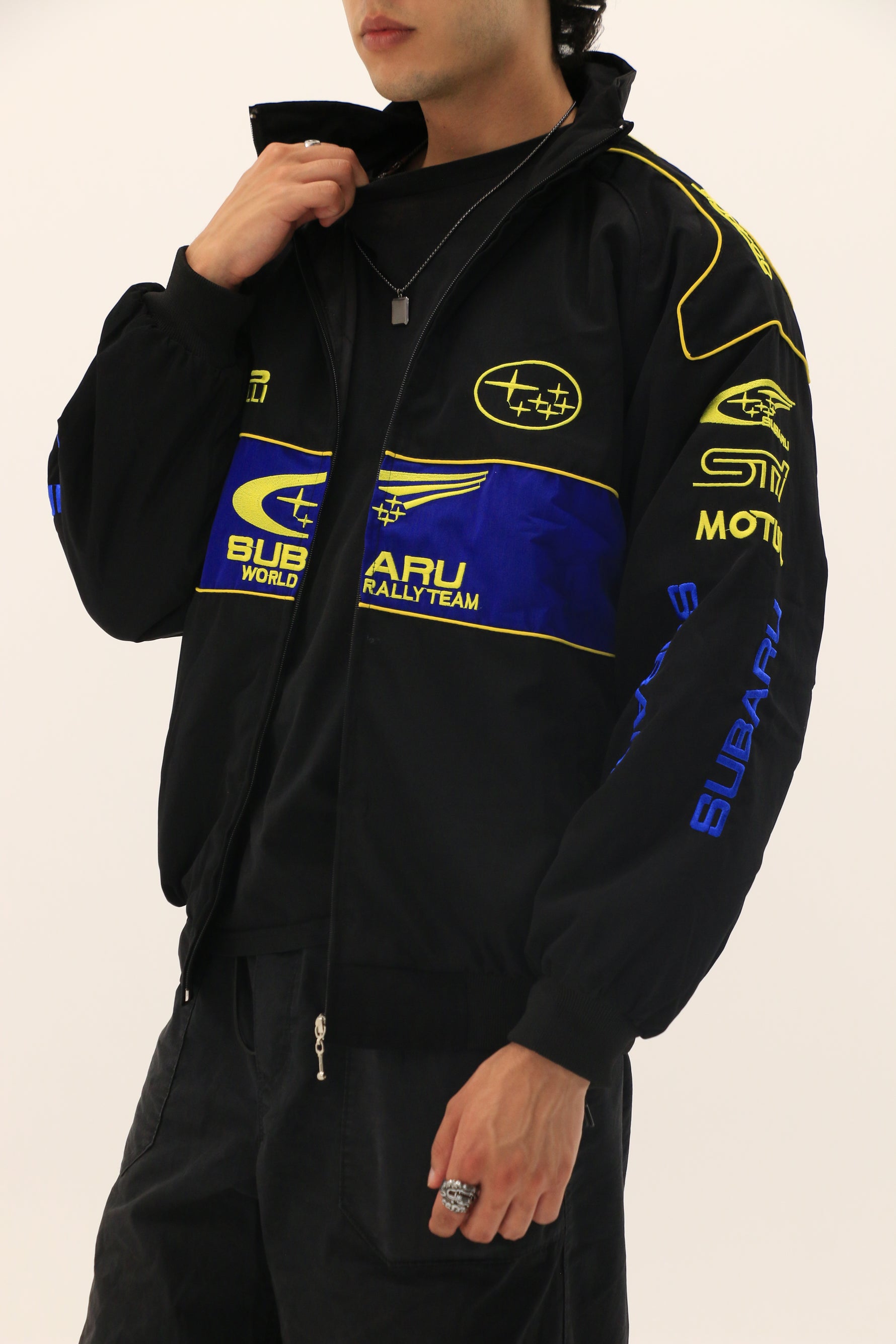 A person with short hair wears a partially unzipped Subaru Rally Vintage Racing Jacket in black with blue and yellow details, paired with black pants, and holds the jacket collar with one hand.