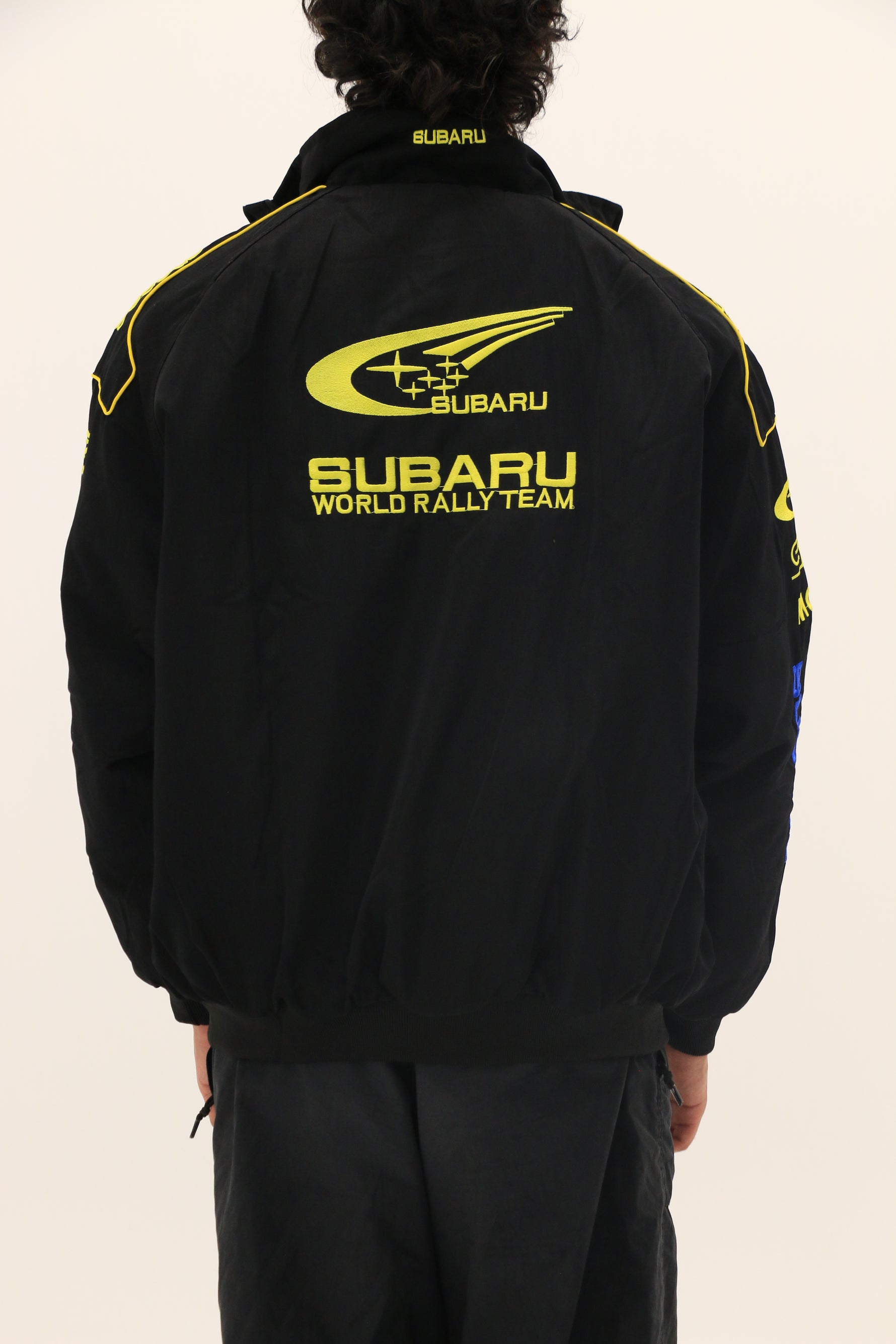 Wearing the Subaru Rally Vintage Racing Jacket—a black piece featuring yellow logos—a person stands with their back to the camera against a plain light background, capturing classic rally heritage.