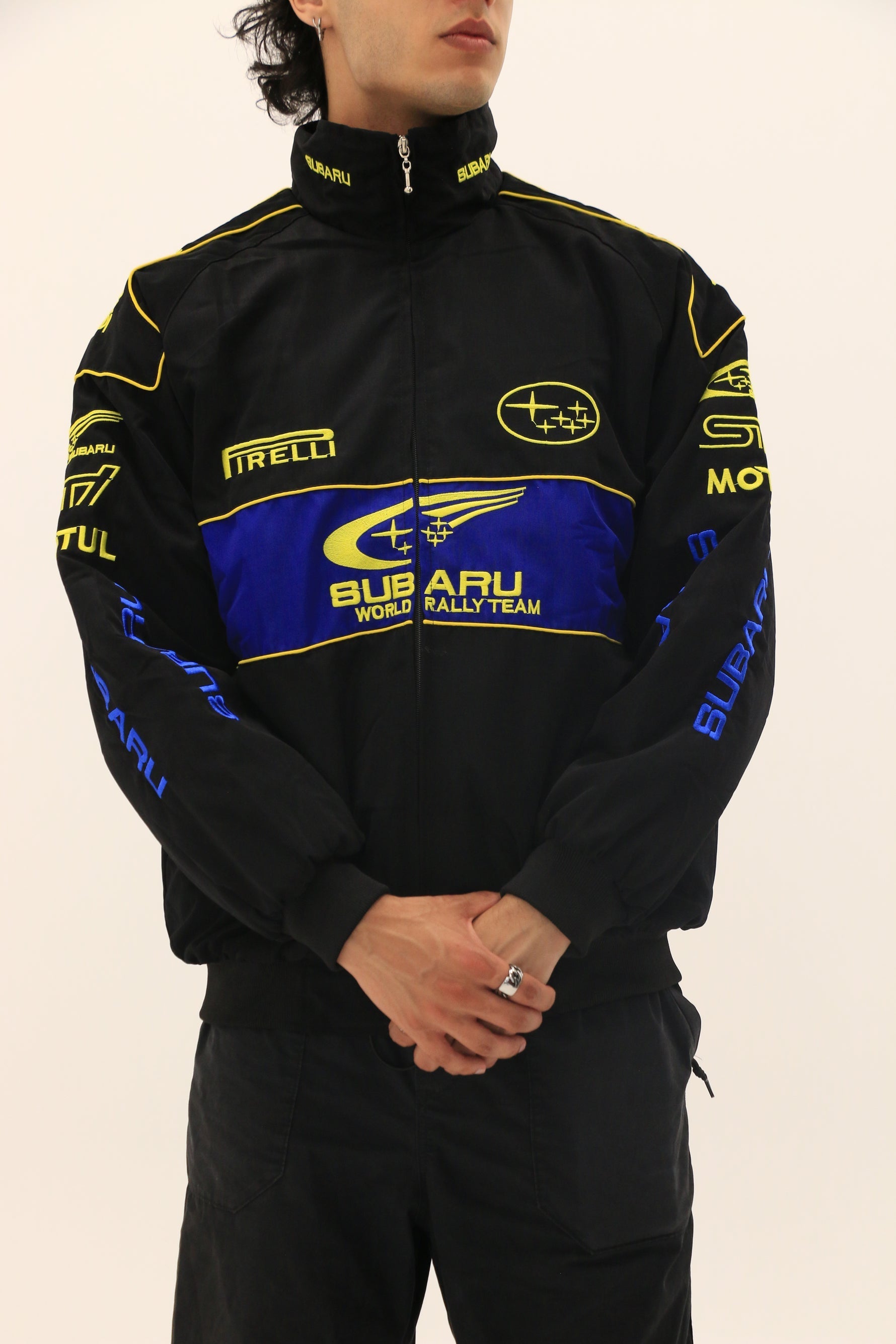 Wearing a Subaru Rally Vintage Racing Jacket in black with yellow and blue accents, a person stands hands clasped; logos such as Subaru, Pirelli, and Motul showcase classic rally heritage.