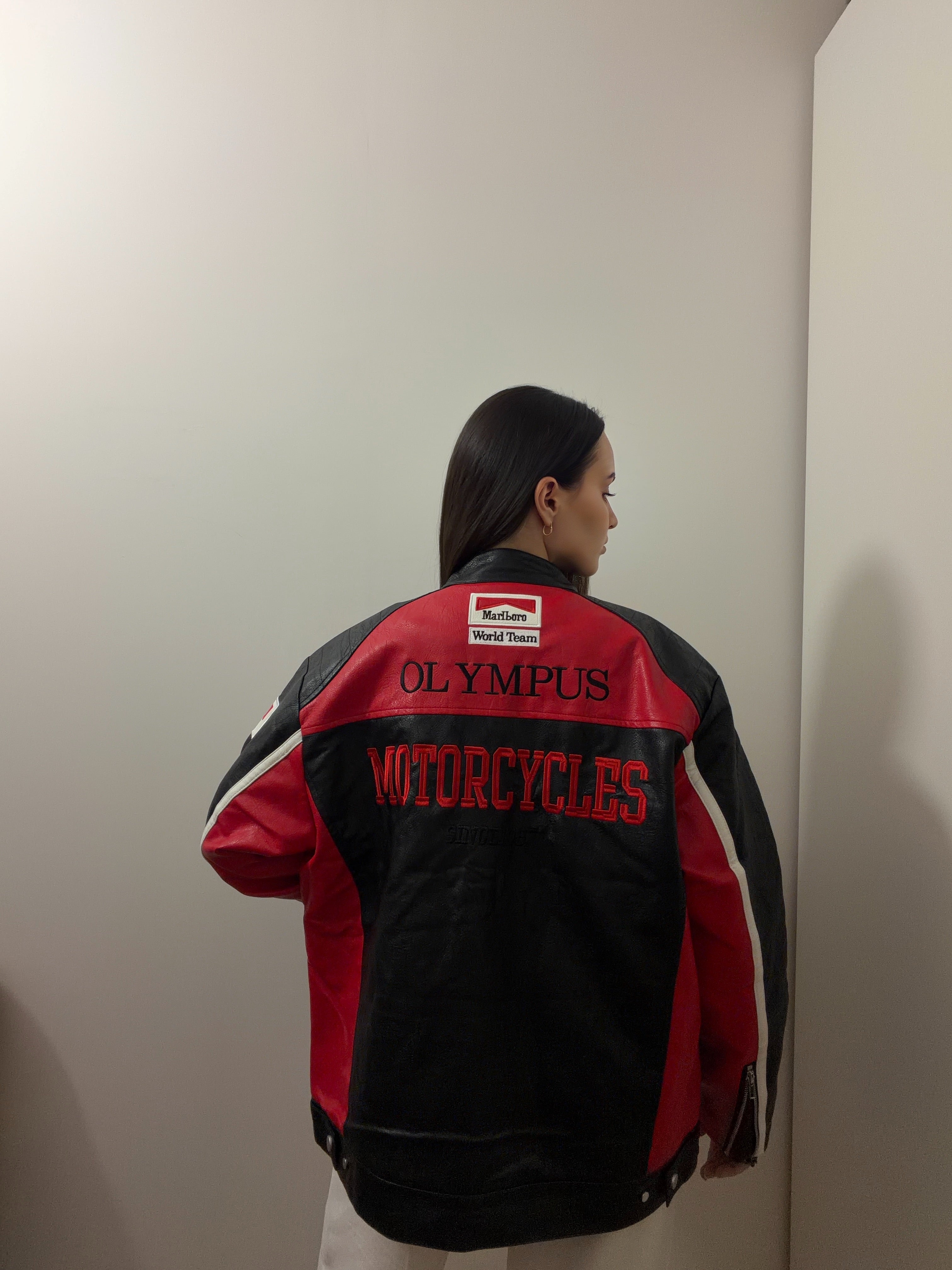 A person with long dark hair faces a wall, wearing a black and red Ferrari Leather Racing Jacket. This motorsport-inspired scene takes place in a plain, indoor room with white walls.