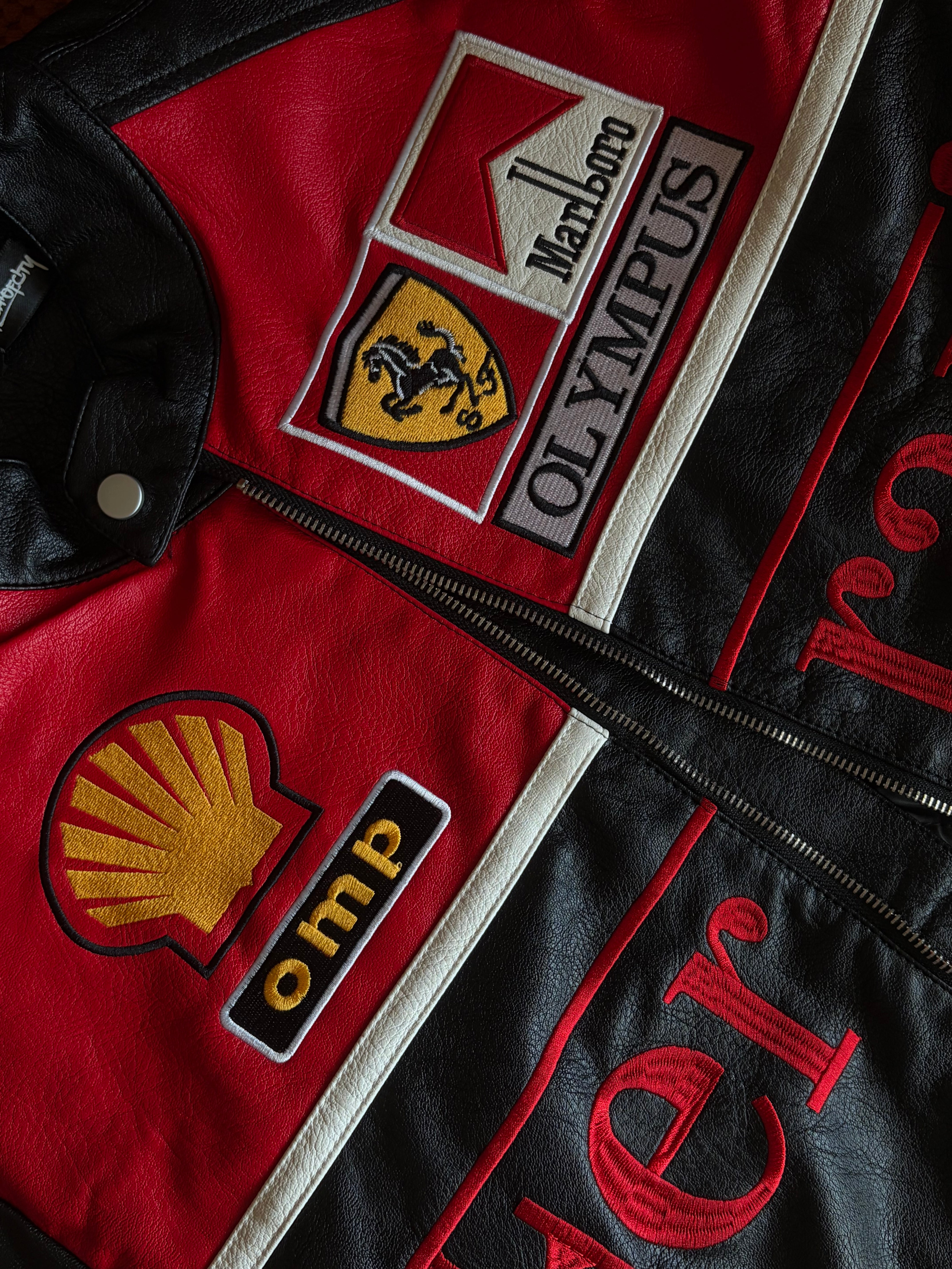 A close-up of the Ferrari Leather Racing Jacket - Black & Red, showcasing Ferrari, Marlboro, Olympus, Shell, and OMP patches. The detailed stitching and vibrant red and black leather emphasize the motorsport-inspired design of this striking jacket.