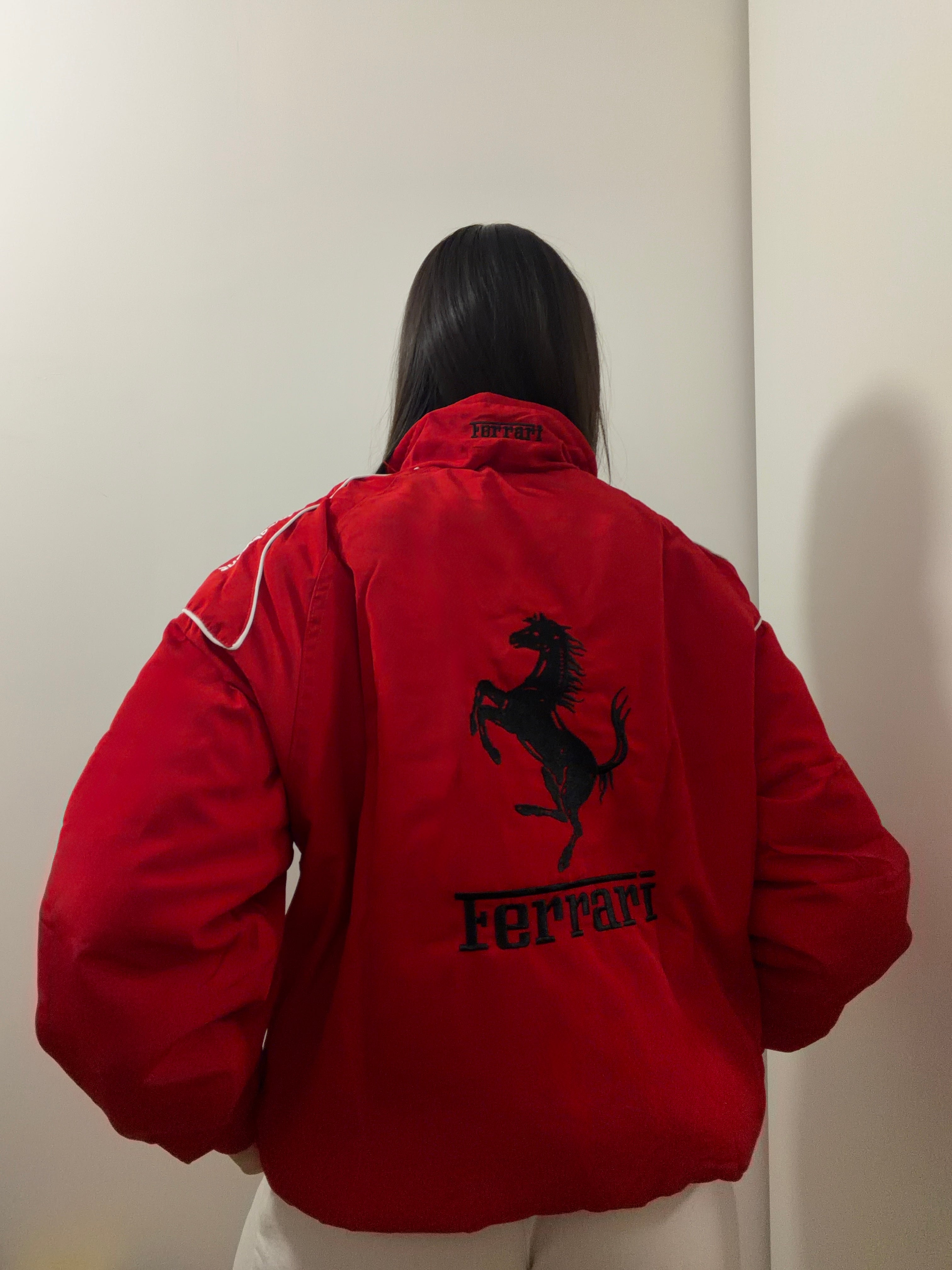 A person with long brown hair, facing away, wears the F1 Vintage Ferrari Jacket - Red Edition, featuring a large black prancing horse logo and Ferrari text on the back, standing against a plain white wall—an iconic look.