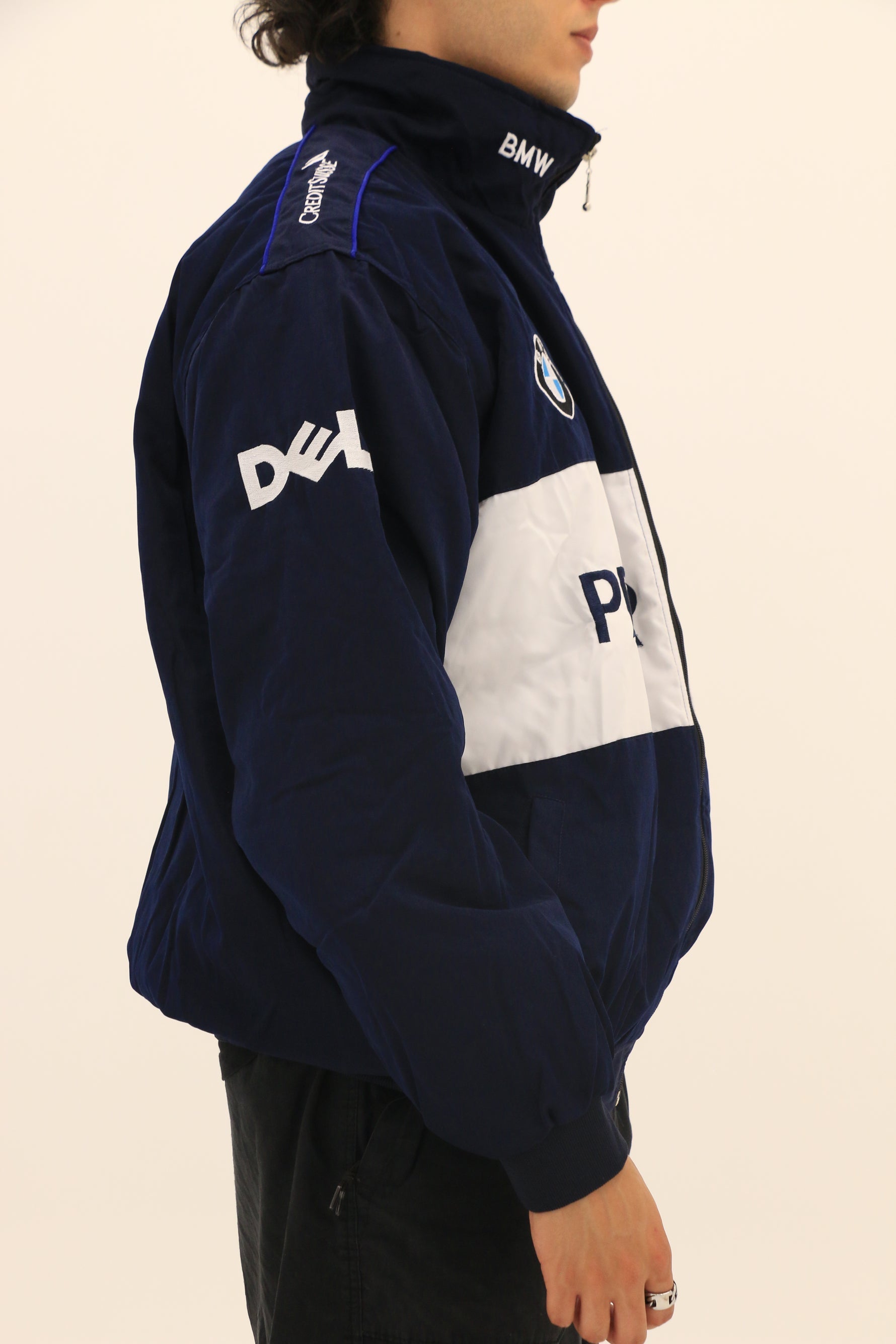 Wearing a BMW F1 Vintage Racing Jacket in navy blue and white with DELL on the sleeve and partially visible POLICE on the front, a person stands against a plain light background, highlighting classic racing heritage.