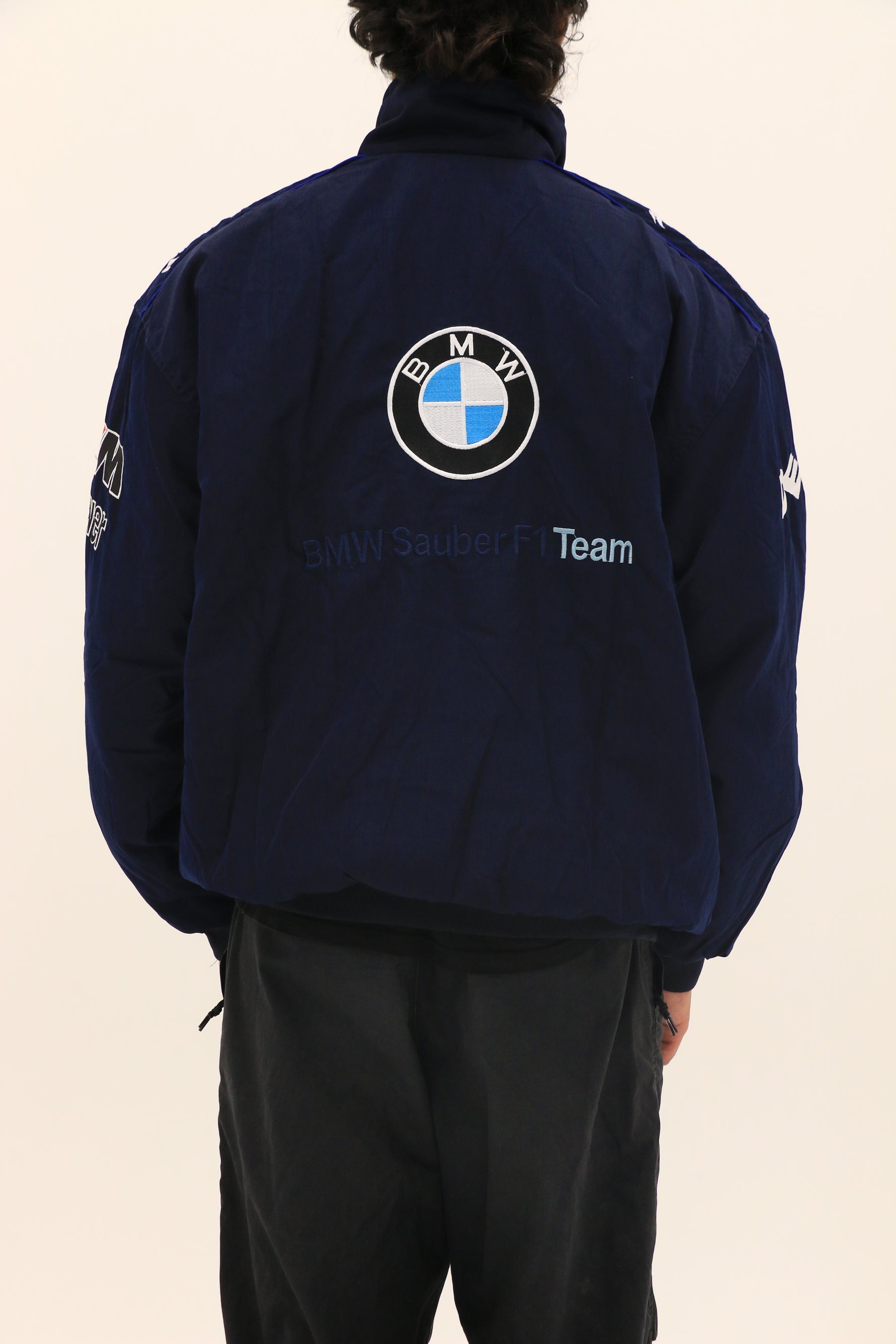 A person stands facing away, wearing a BMW F1 Vintage Racing Jacket in navy blue with a large BMW logo and BMW Sauber F1 Team text on the back, plus sleeve logos. The plain, light background accentuates the jacket’s classic racing style.