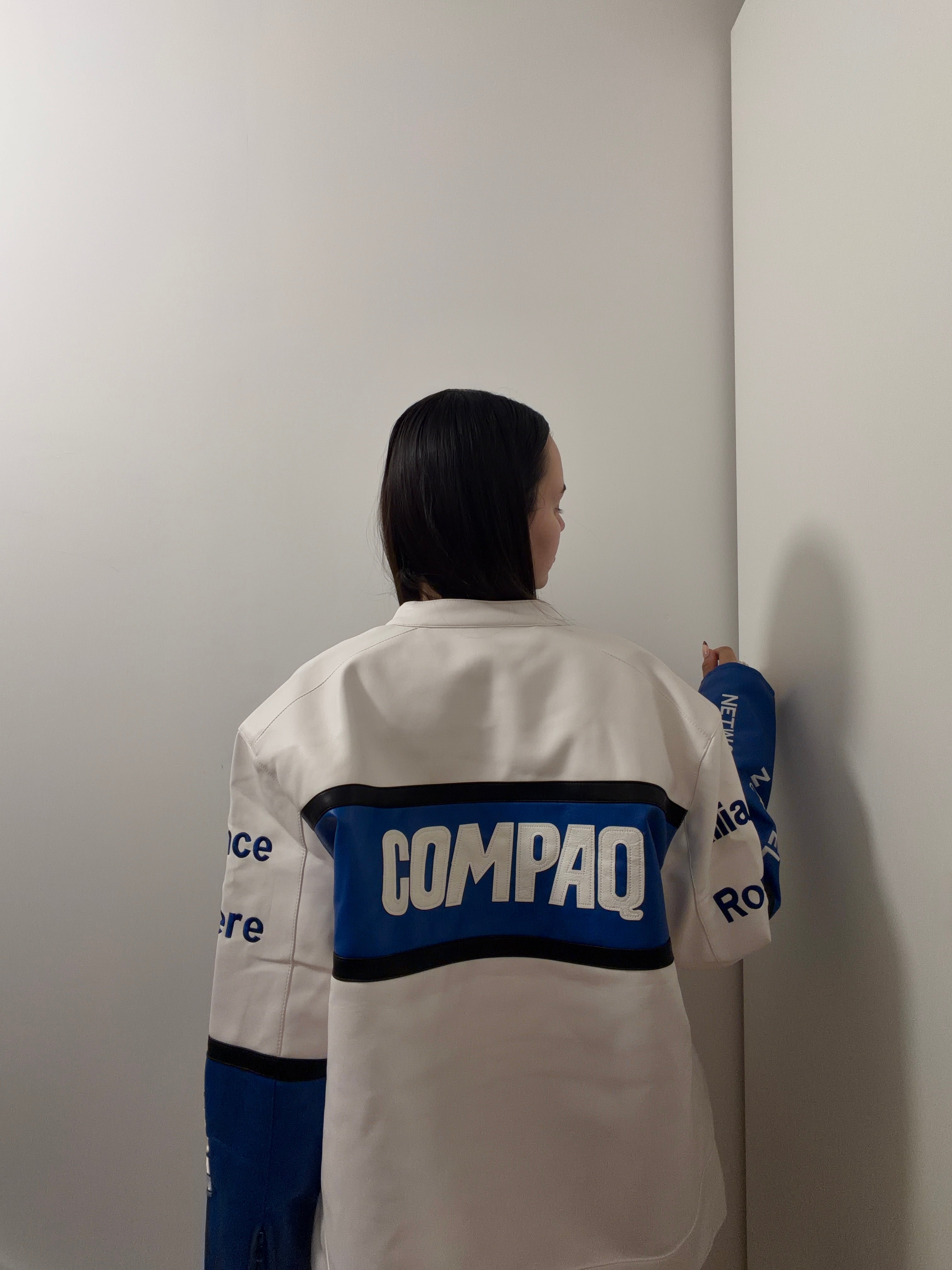 A person with long dark hair, seen from behind, stands in a corner wearing the BMW Vintage Leather Jacket—a white and blue piece with a racing-inspired design that hints at motorsport heritage.