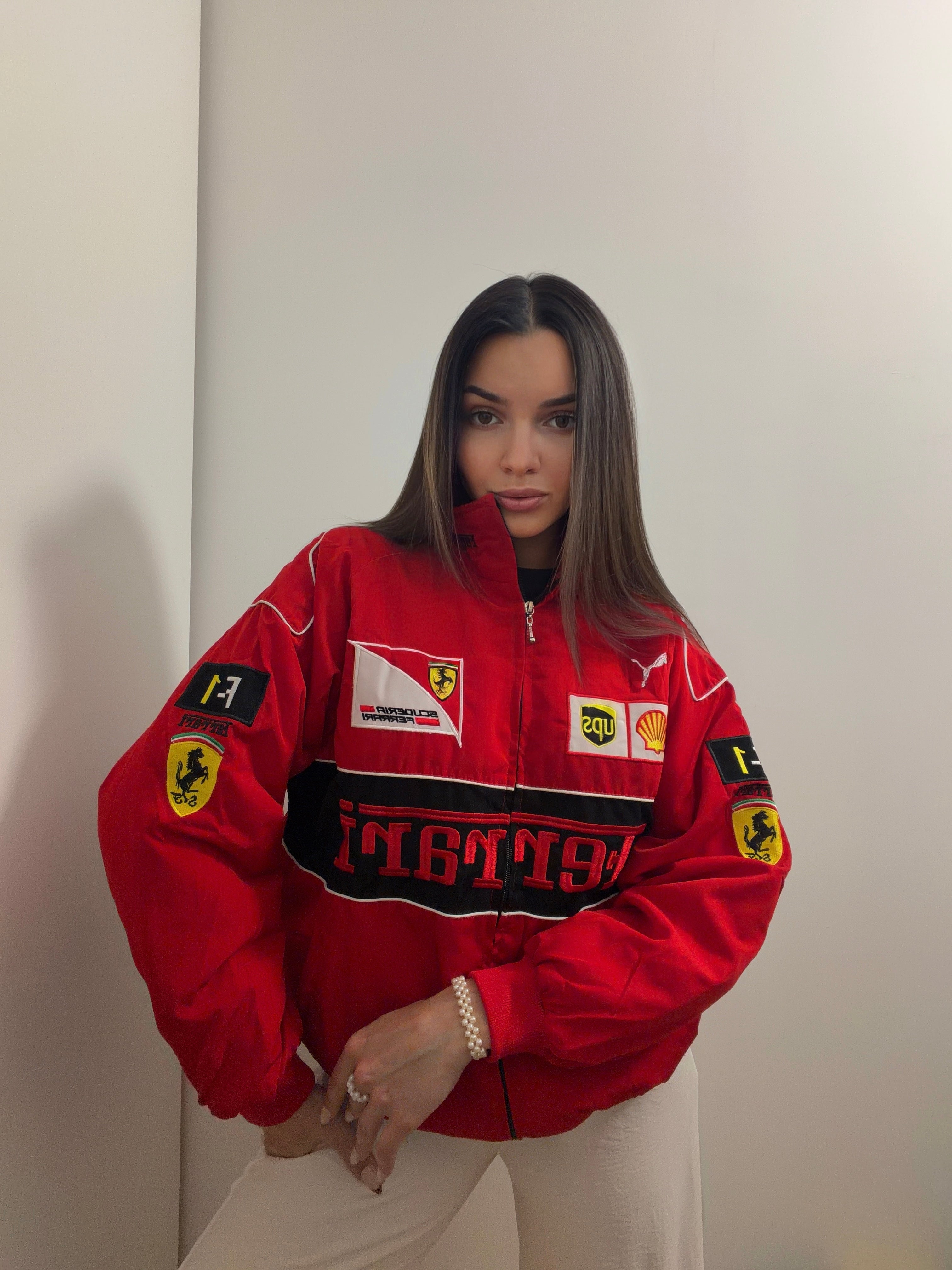 A woman with straight brown hair stands indoors against a plain white wall, wearing the F1 Vintage Ferrari Jacket - Red Edition emblazoned with sponsor logos, channeling the essence of classic Ferrari racing style.