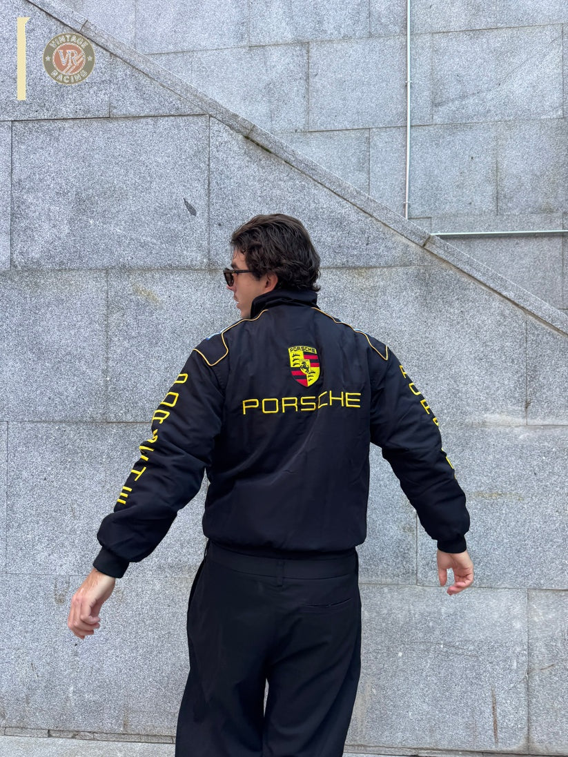 A person wearing a Porsche Vintage Racing Jacket with yellow lettering and logo stands outdoors, facing away by a gray stone wall with steps and railings, capturing the essence of true motorsport heritage.