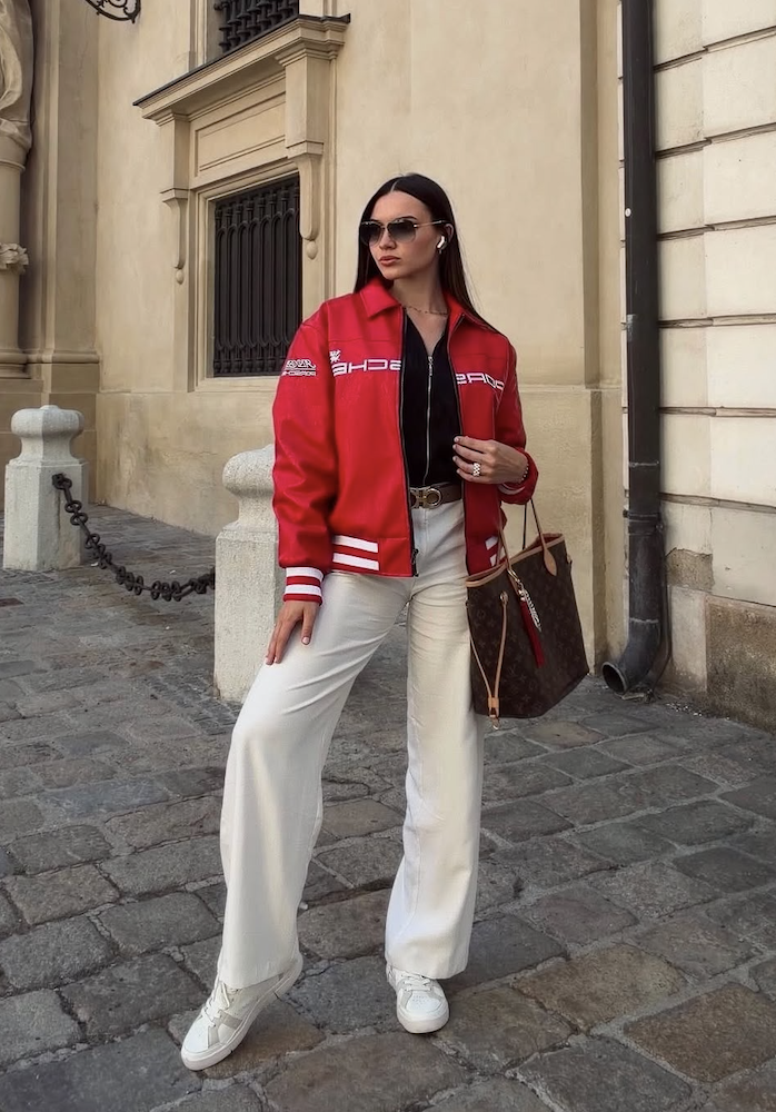 A woman wearing the Vintage Porsche Leather Jacket - Red Edition, paired with white pants and white sneakers, stands on a cobblestone street holding a brown handbag and wearing sunglasses.