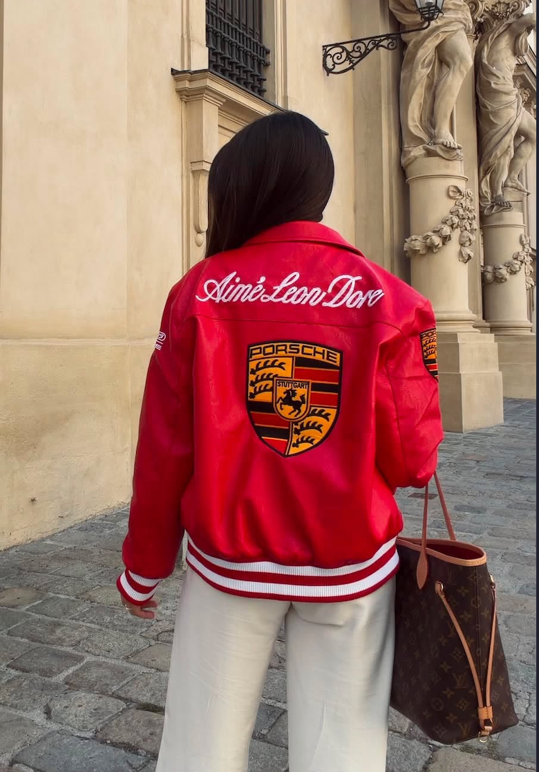 A person wearing a Vintage Porsche Leather Jacket - Red Edition by Aimé Leon Dore and white pants stands on a cobblestone street, holding a brown Louis Vuitton bag and facing away from the camera.