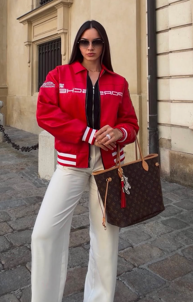 A woman poses on a cobblestone street wearing the Vintage Porsche Leather Jacket - Red Edition, paired with white pants and sunglasses, while holding a Louis Vuitton tote bag adorned with a scarf. She stands in front of a beige building.
