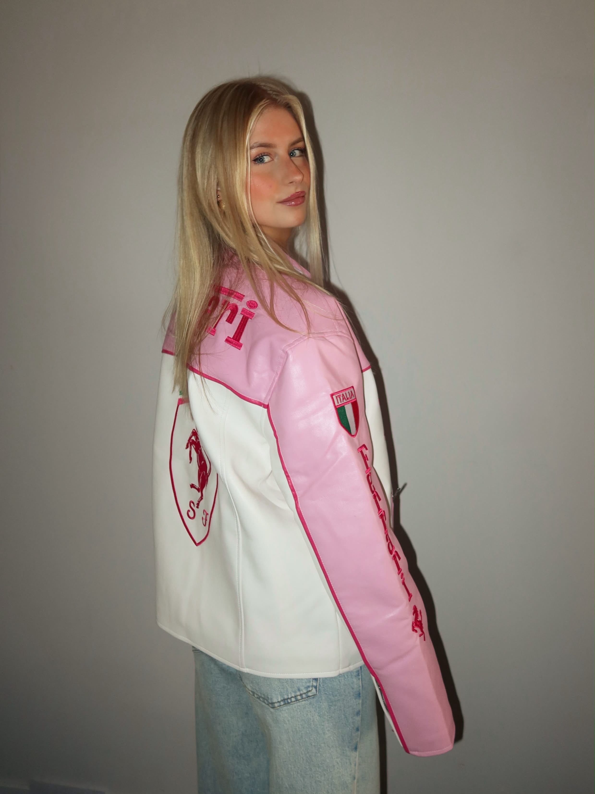 A woman with long blonde hair stands sideways and looks over her shoulder in front of a plain light-colored wall, wearing the Ferrari F1 Vintage Racing Leather Jacket – Pink Edition, which features motorsport-inspired patches.