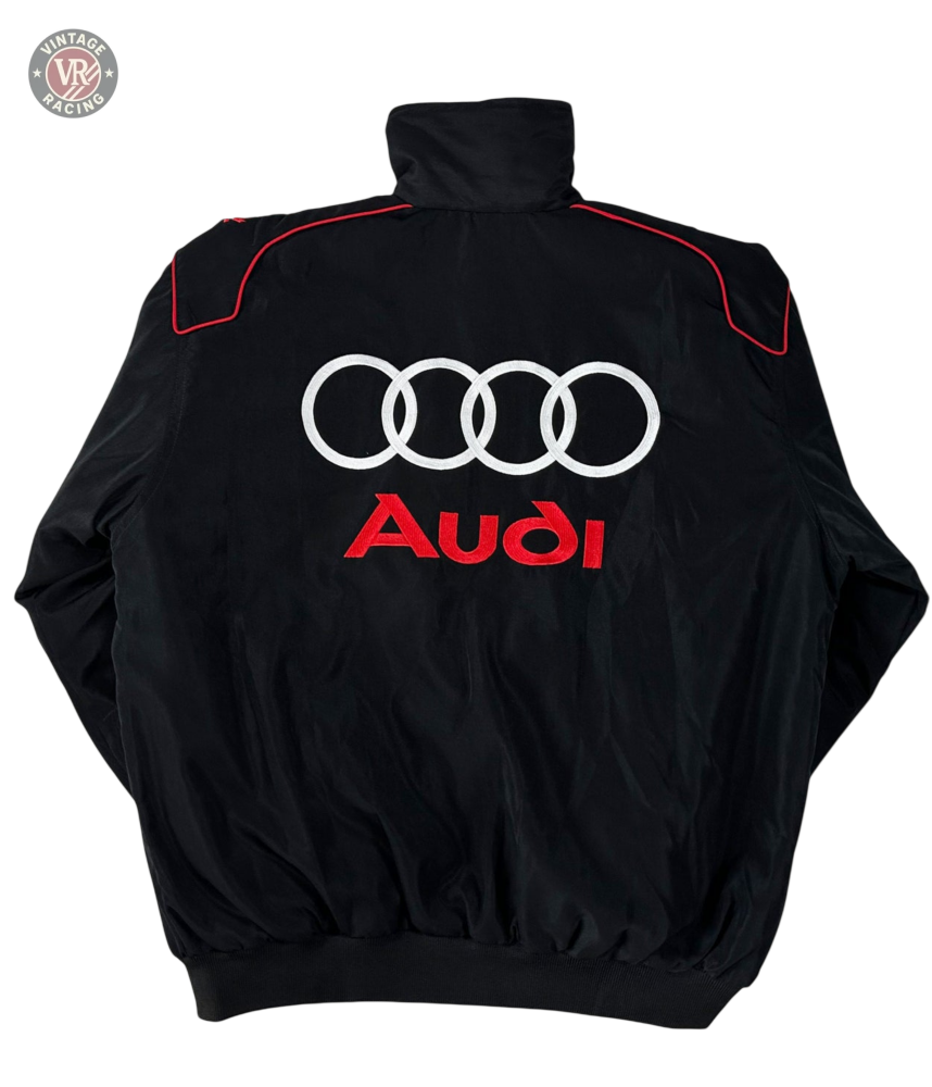 The Audi Vintage Racing Jacket is black with four white interlocking rings and red Audi embroidery on the back. Featuring a high collar, red shoulder accents, and vintage motorsport style, it celebrates Quattro heritage.