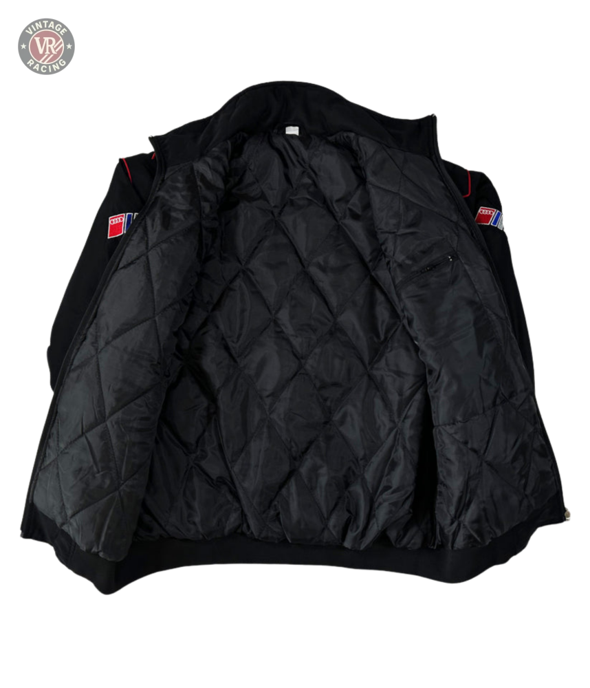 The Audi Vintage Racing Jacket is black with red and blue sleeve patches, features a quilted inner lining, and has a small zippered pocket on the left side—capturing classic vintage motorsport style.