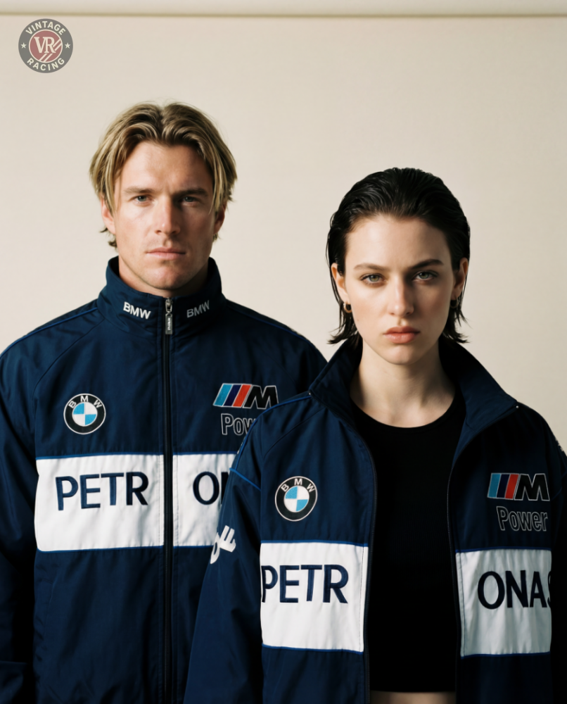 A man and woman wear matching navy blue BMW F1 Vintage Racing Jackets with white stripes and PETR and ONAS detailing, posing against a plain backdrop, capturing authentic racing heritage.