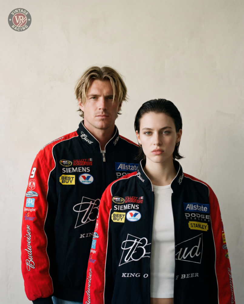A man and woman stand side by side against a plain light background, both wearing the Budweiser Vintage Racing Jacket - NASCAR Black Edition with colorful sponsor patches, looking directly at the camera with serious expressions.