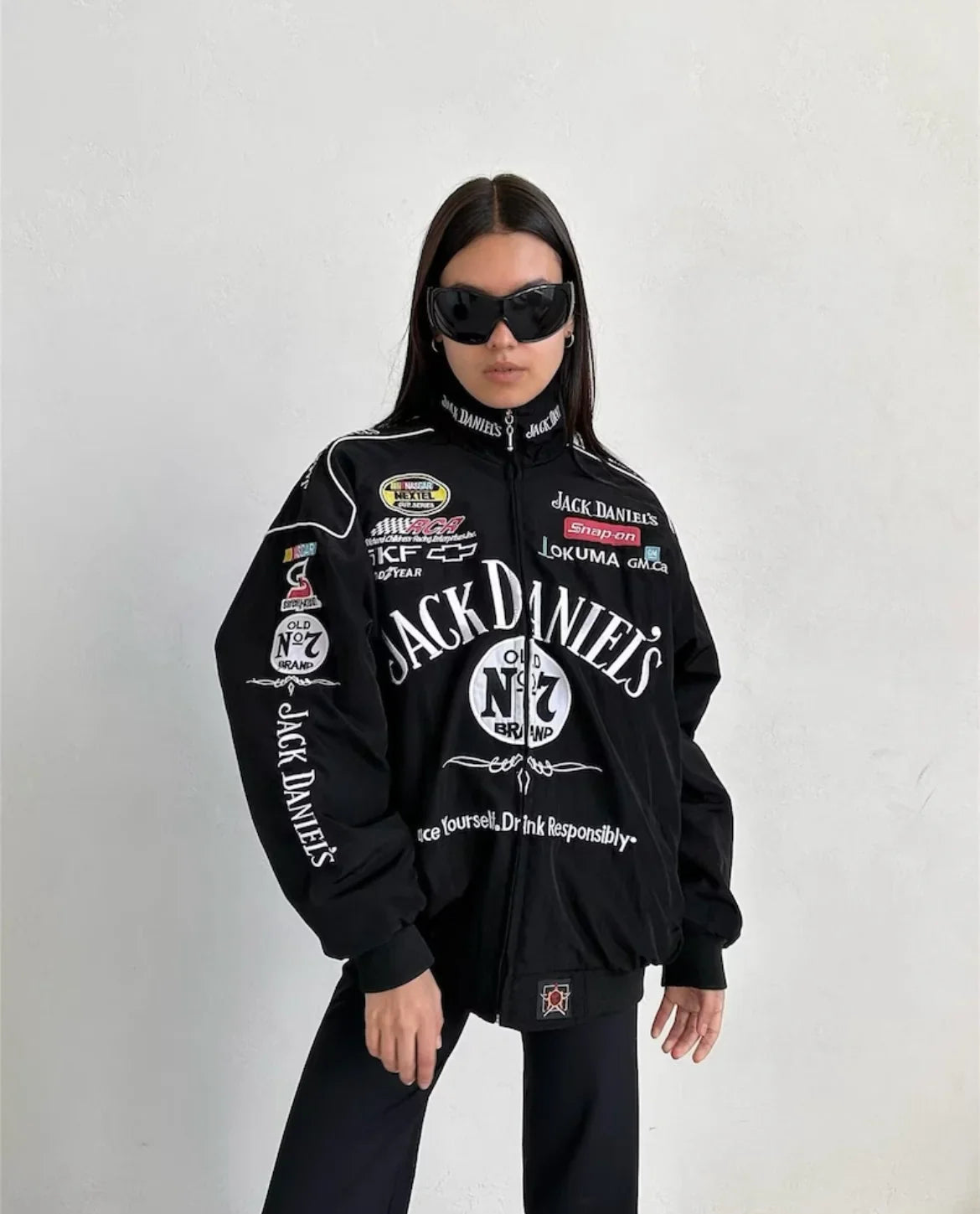 A woman in black sunglasses and a Jack Daniel’s Racing Vintage Jacket stands against a white wall with her hands in her pockets, showcasing confident, racing-inspired streetwear style.