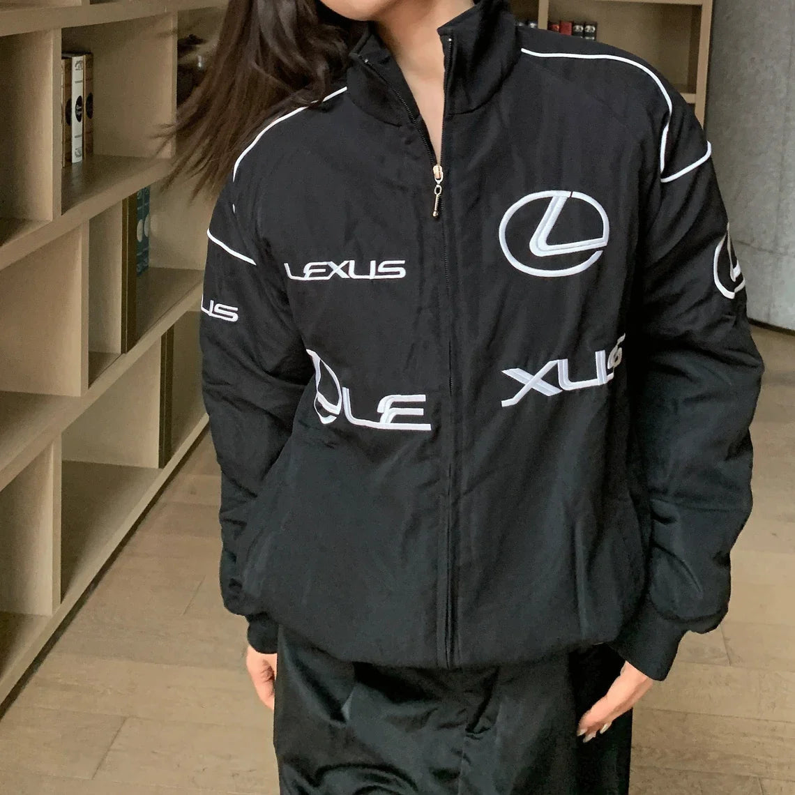 A person models the Lexus Vintage Racing Jacket—black with bold white logos and lettering—indoors by bookshelves, embodying luxury motorsport style with a partially turned head and hair obscuring part of their face.