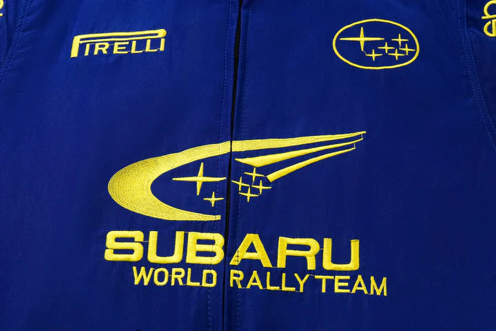 The Subaru World Rally Team Vintage Jacket is a blue fabric jacket with yellow embroidery featuring the team’s swoosh-and-stars logo, Pirelli branding, and Subaru logos inspired by the iconic 555 livery.