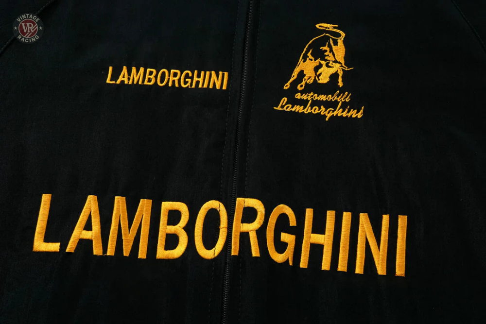 Close-up of the Lamborghini Vintage Racing Jacket, a black retro jacket with yellow LAMBORGHINI embroidery, the classic bull logo, and “automobili Lamborghini”—ideal for motorsport fans.