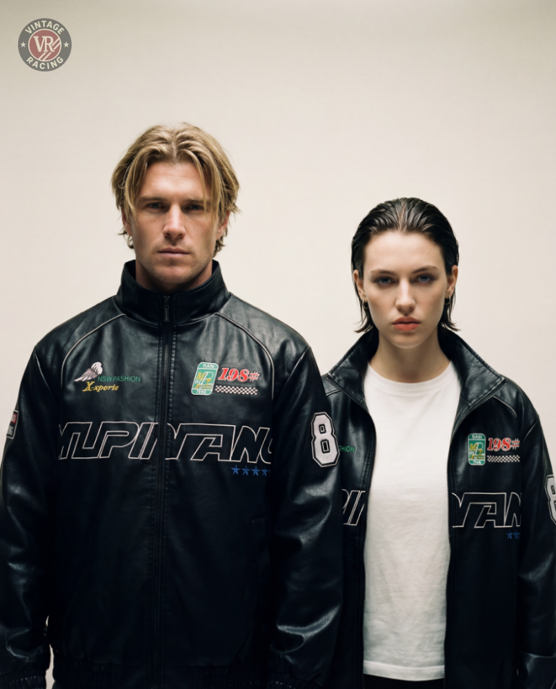 A man and a woman stand side by side against a plain background, both wearing the LAP 85 Black Racing Jacket with white text and patches. The woman pairs hers with a white shirt, embodying F1-inspired style with neutral expressions.